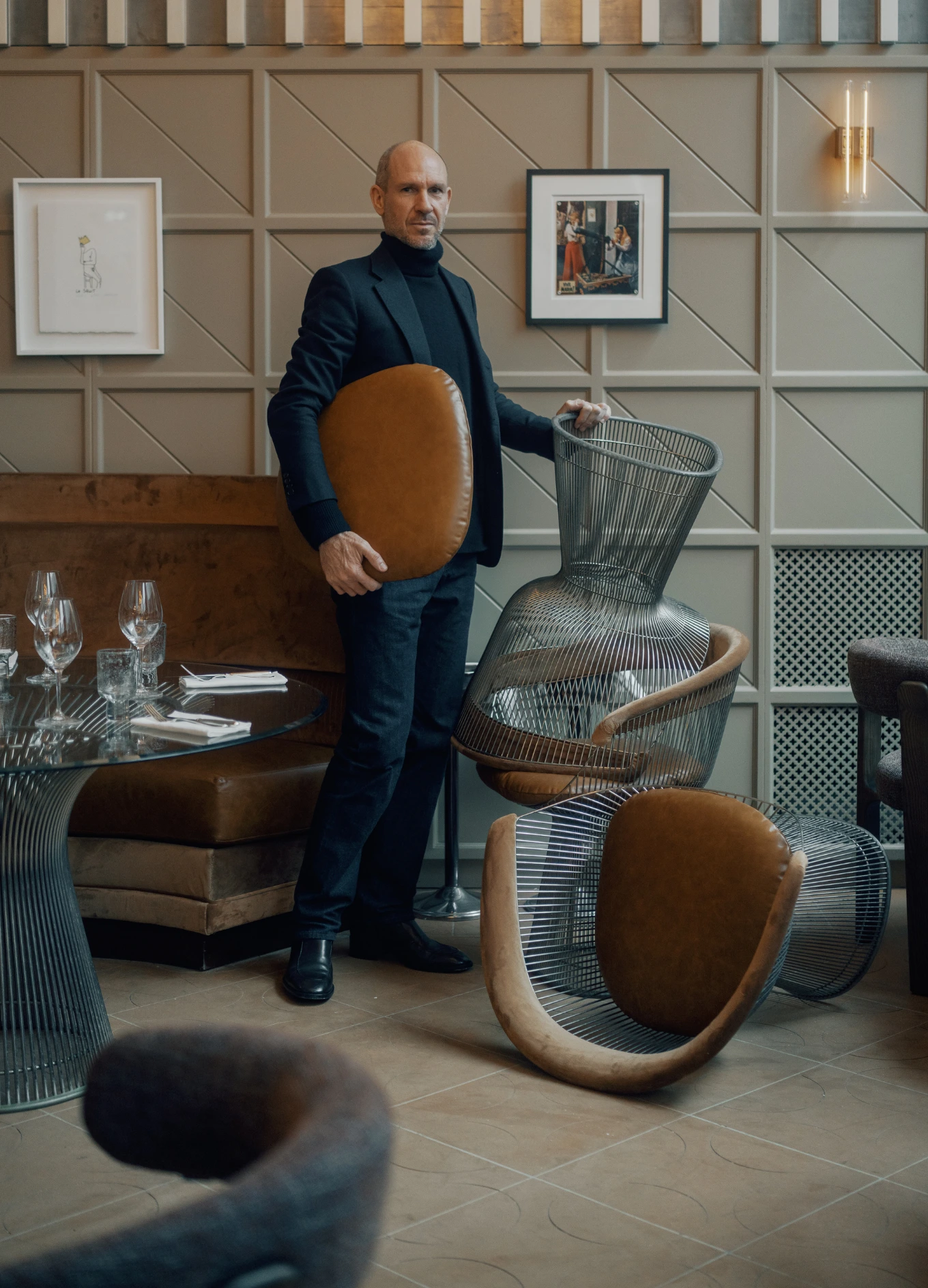 Man in black turtleneck and blazer holding a brown chair cushion while standing next to modern wire-frame chairs inside a stylish restaurant.