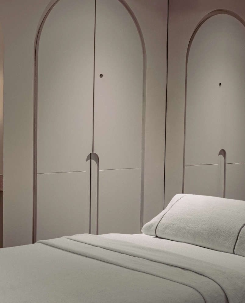 Minimalist bedroom with a neatly made bed featuring a white pillow and blanket, and light-colored arched closet doors in the background.