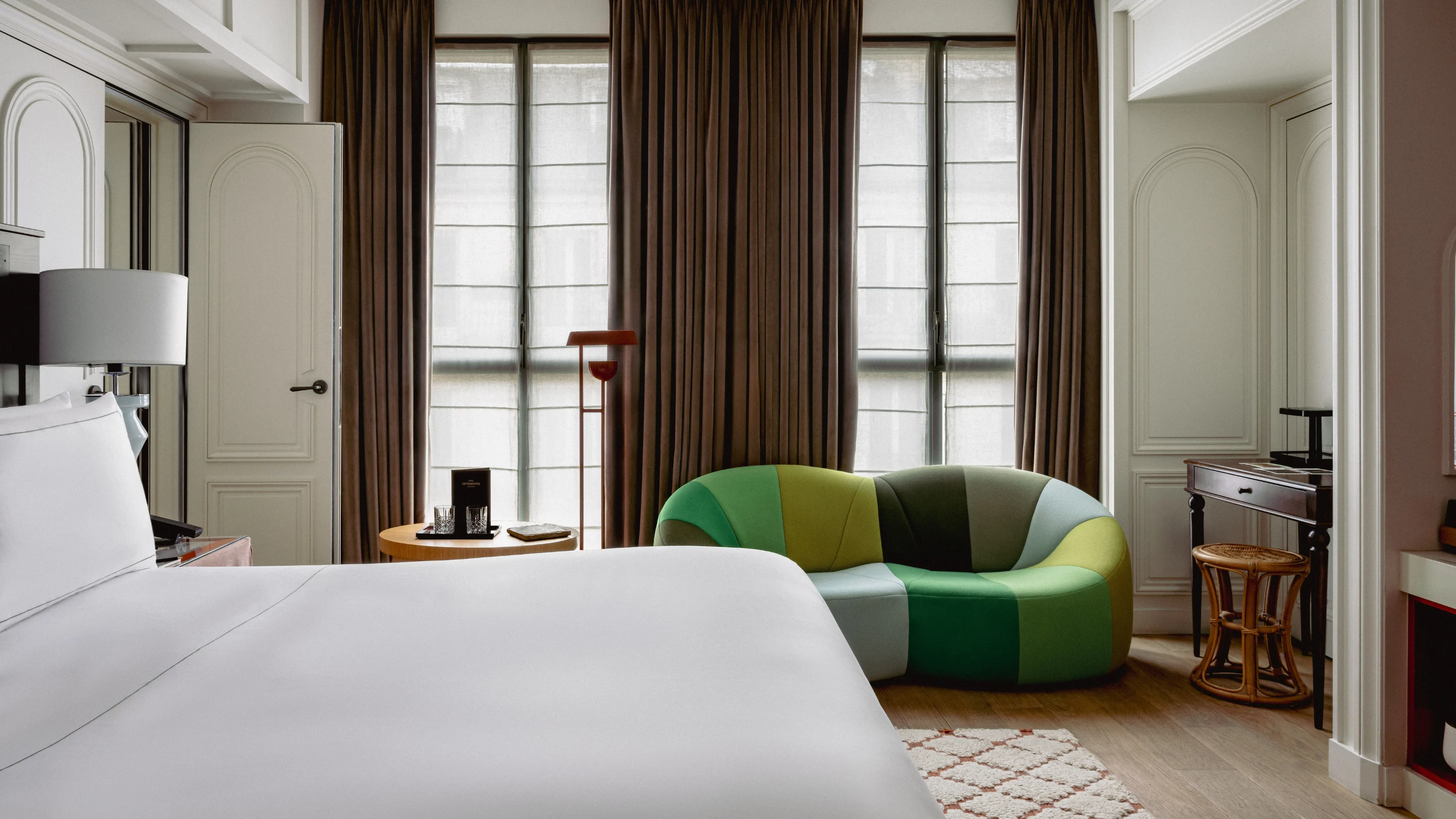Modern hotel room with white bed, multicolored green sofa, round wooden table, floor lamp, and large windows with brown curtains.