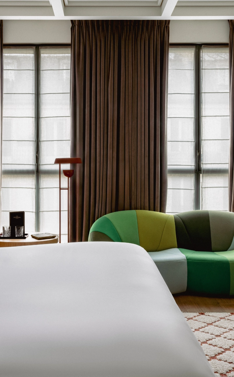 Hotel room with white bedspread, colorful patchwork curved sofa, brown floor lamp, round wooden side table, and large windows with sheer and dark curtains.