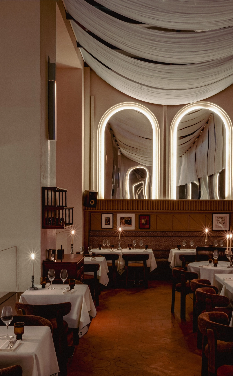 Dimly lit upscale restaurant interior with white tablecloths, cushioned chairs, candles on tables, and large illuminated arched mirrors with draped ceiling fabric.