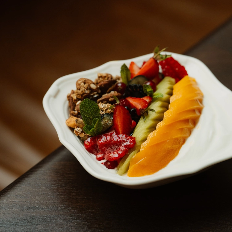 Bowl of sliced mango, kiwi, strawberries, raspberries, granola, and mint leaves on a dark wooden surface.