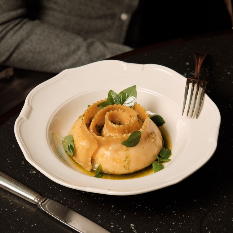 Plate of rolled pasta resembling a flower with green herb garnish and olive oil on a white dish, accompanied by a knife and fork on a dark table.