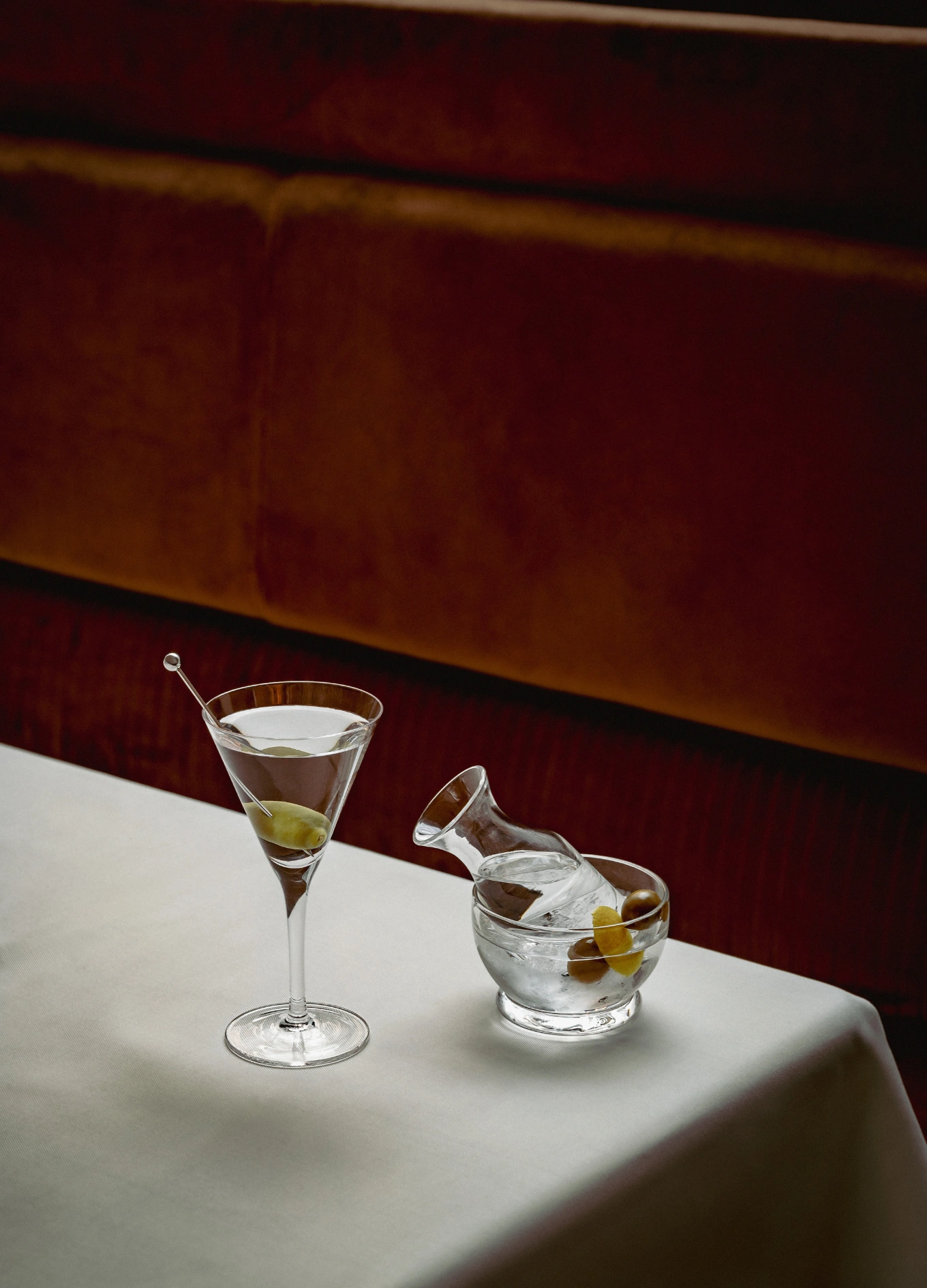 Martini glass with an olive on a pick and a small carafe in a bowl of ice with olives on a white tablecloth.