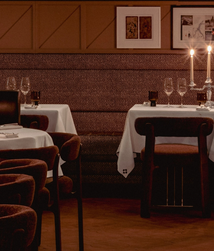 Cozy restaurant interior with tables covered in white tablecloths, brown upholstered chairs, candlesticks, and framed artwork on the wall.