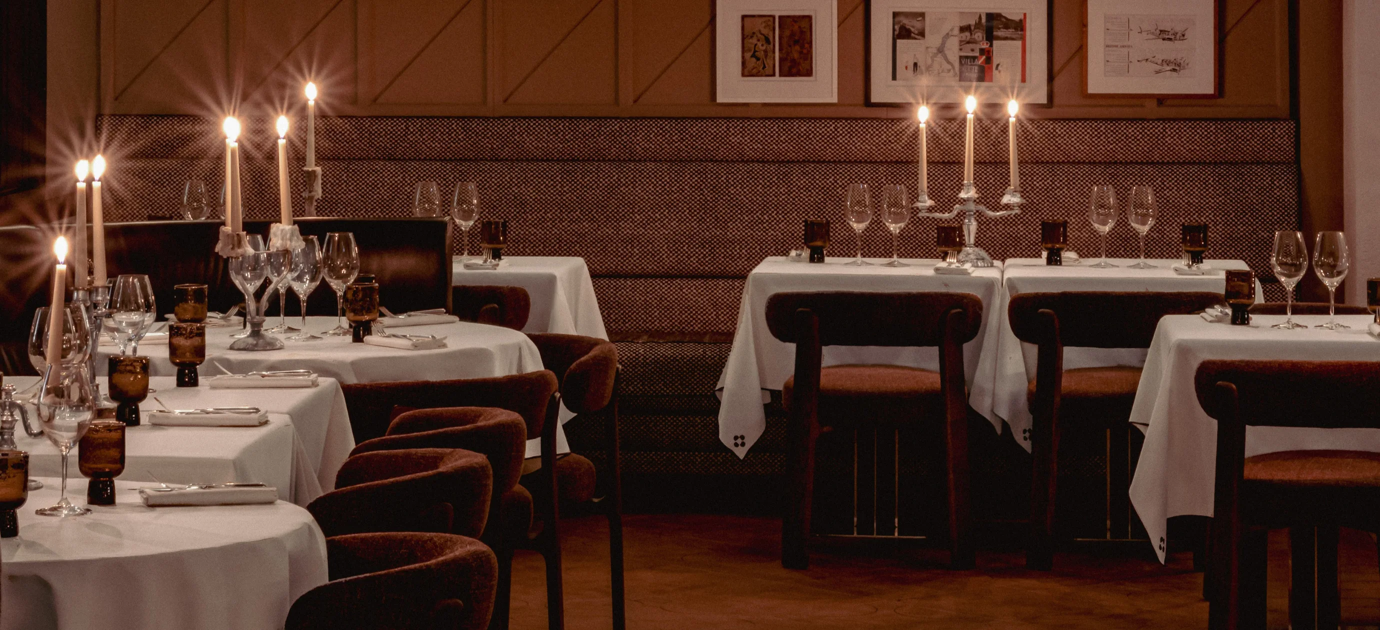Elegant restaurant dining area with tables set with white tablecloths, wine glasses, brown glassware, and lit candles in candelabras.