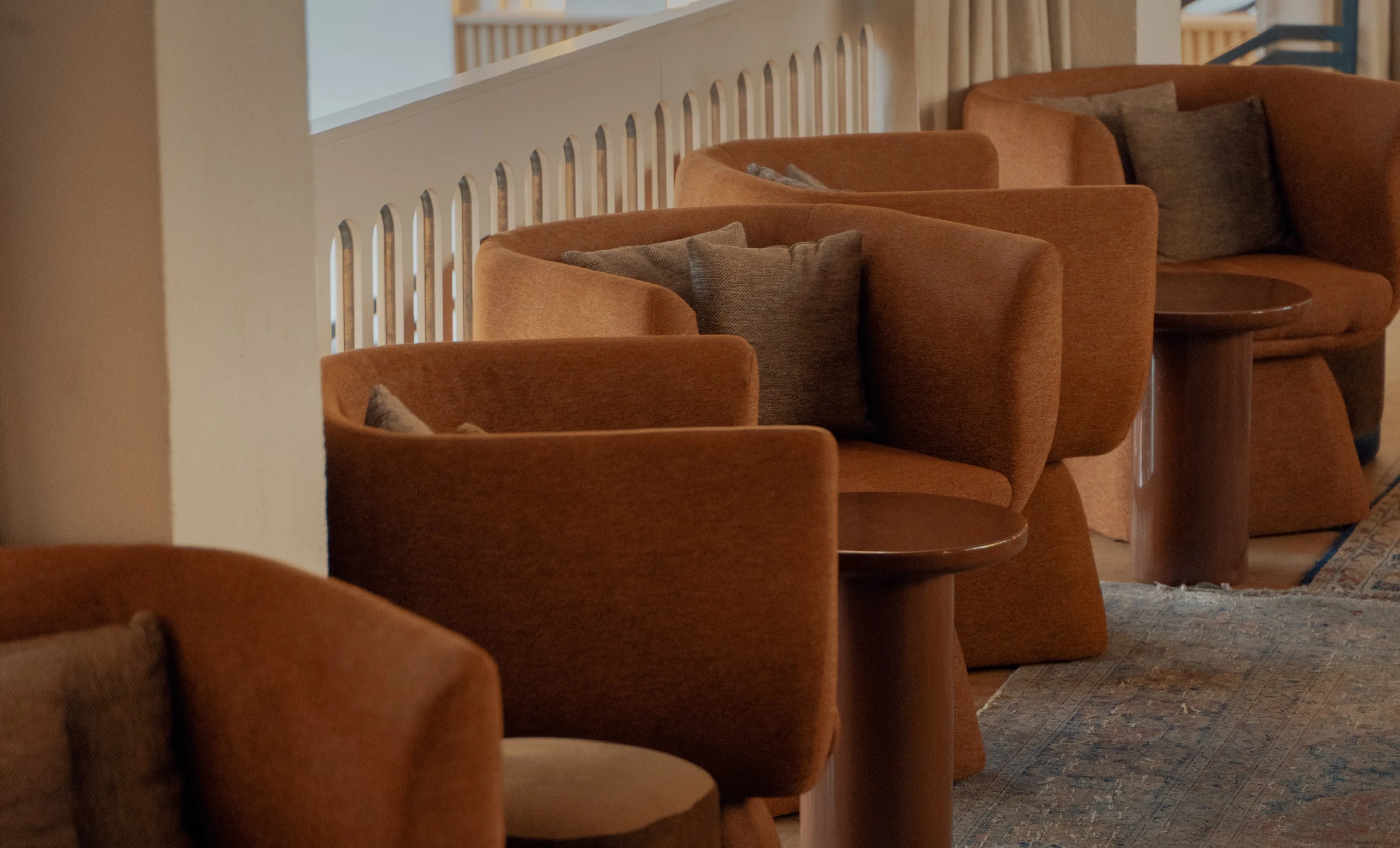 Row of brown upholstered armchairs with gray cushions next to small round wooden tables on a patterned carpet.