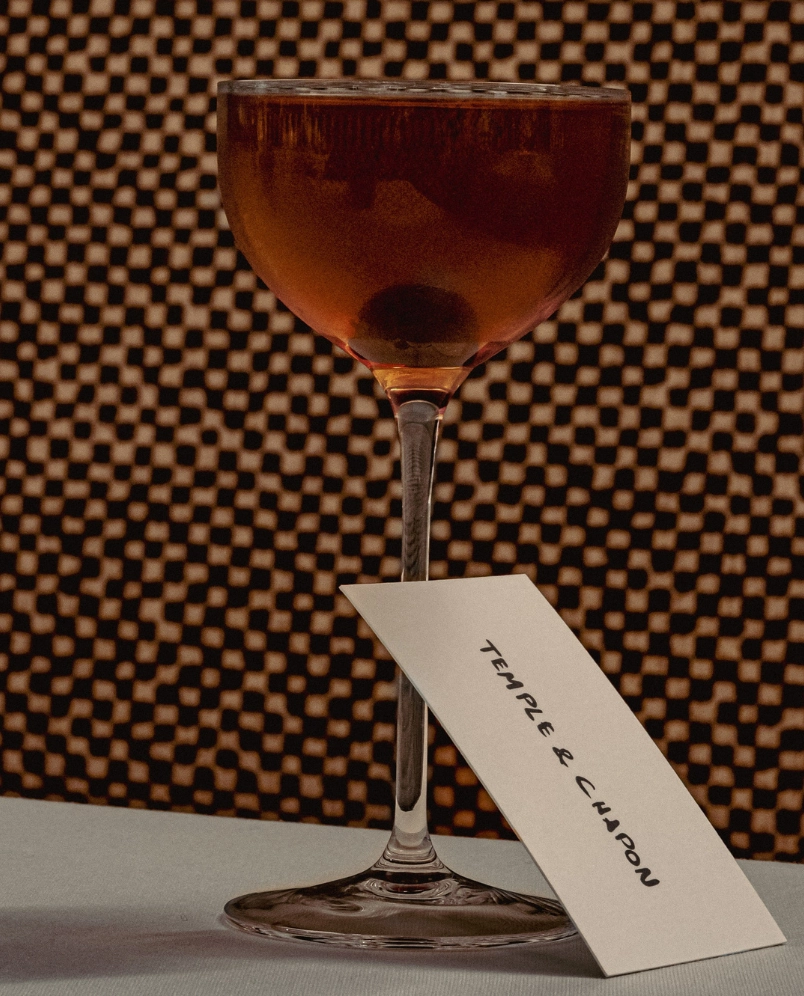 Cocktail glass filled with a dark amber drink with a cherry at the bottom and a card reading 'TEMPLE & CHAPON' leaning against the stem.