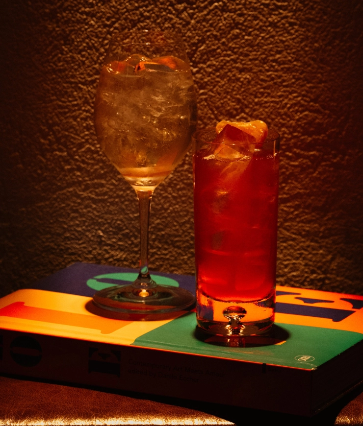 Two cocktail glasses with ice on a colorful book against a textured brown wall under warm lighting.