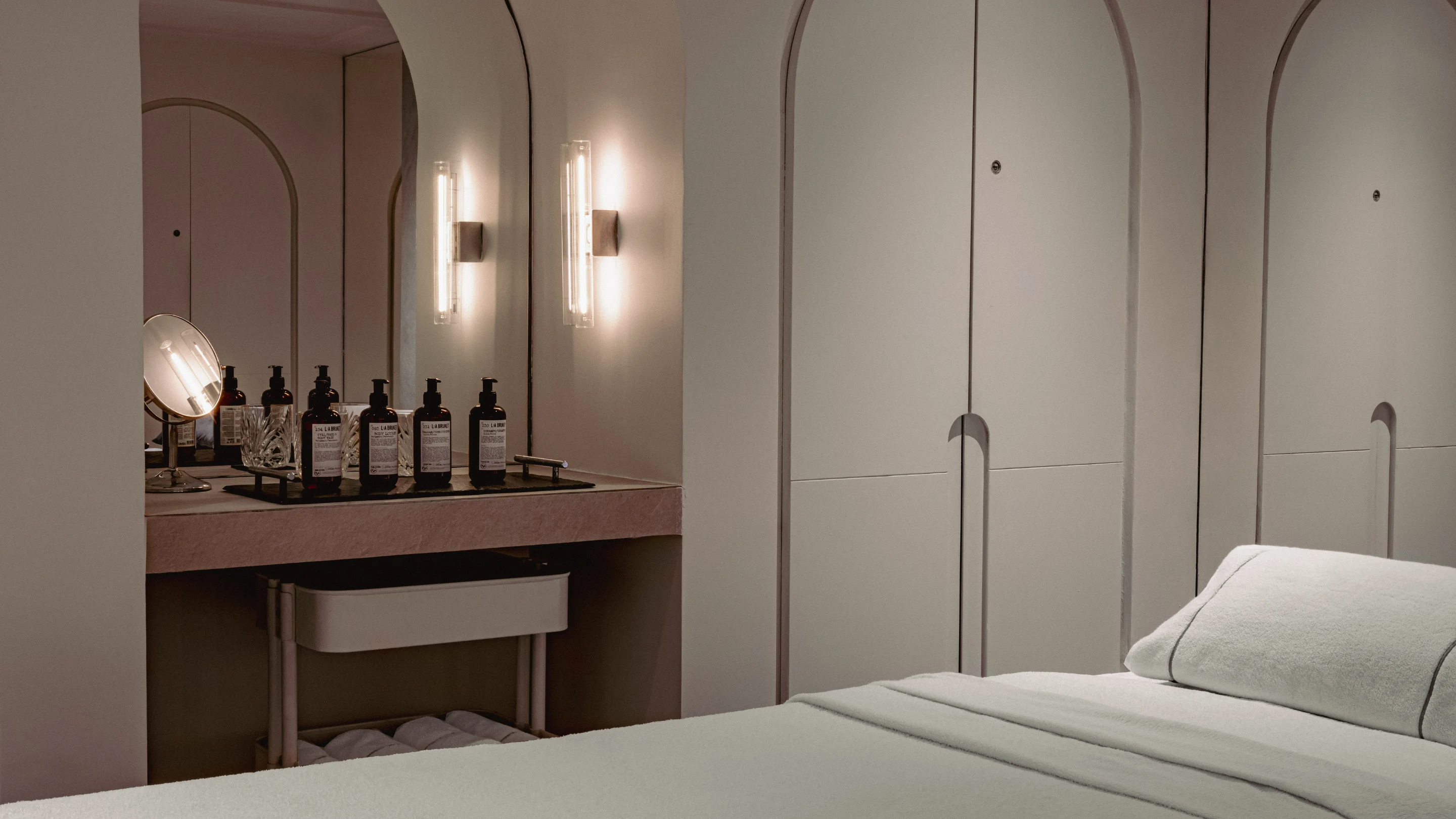 Minimalist spa treatment room with a massage bed, towel, skincare bottles on a tray, and soft wall lighting.