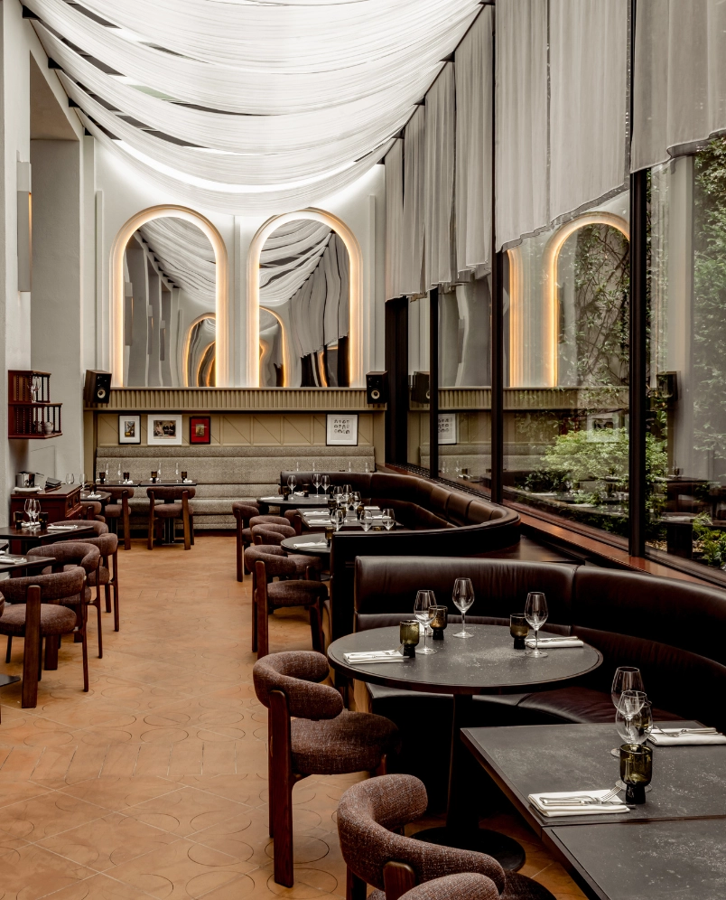 Elegante interior de restaurante con mesas redondas y rectangulares, sillas lujosas, grandes ventanales con cortinas y arcos decorativos con iluminación.