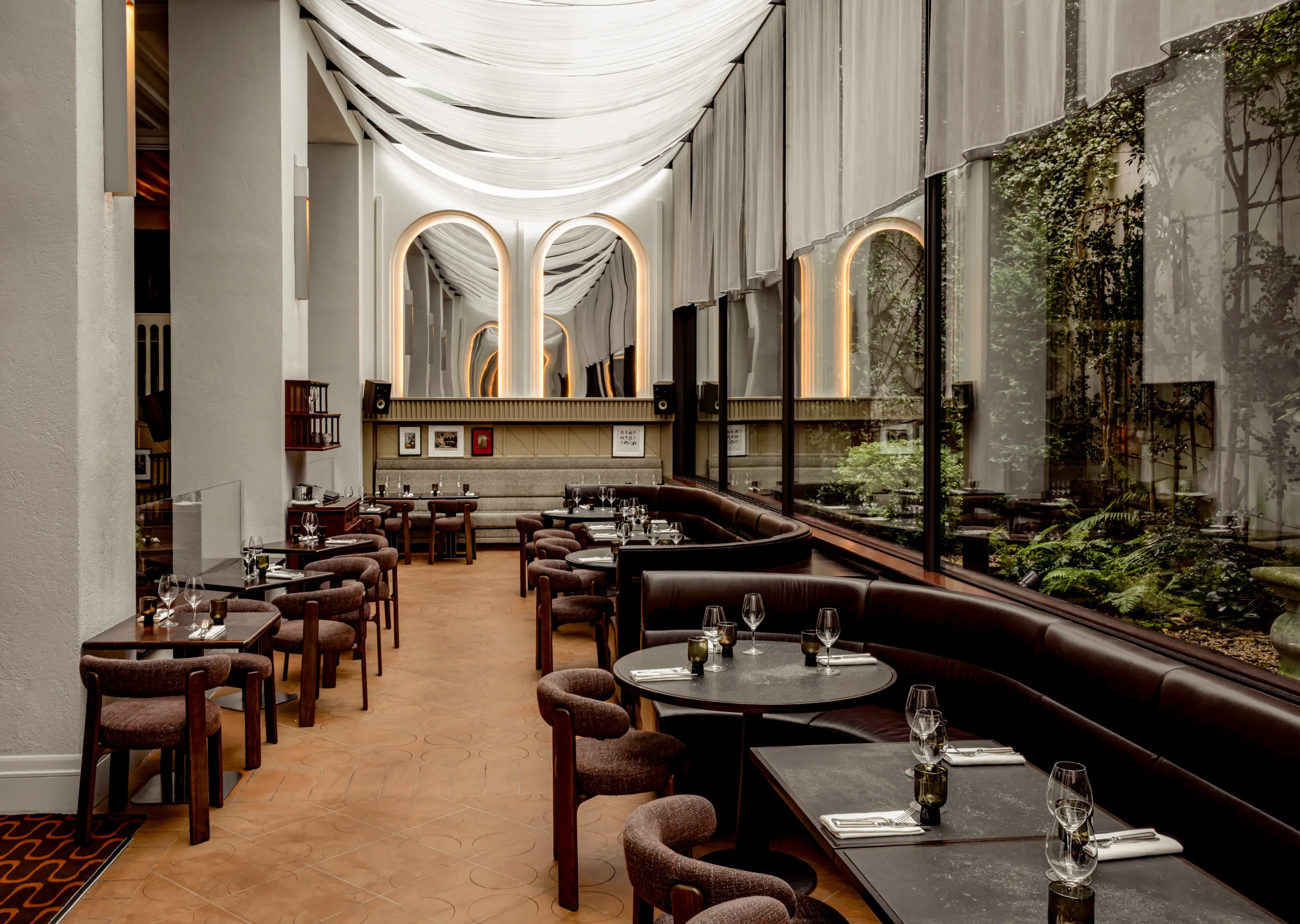 Área de descanso interior del restaurante con mesas redondas y cuadradas, sillas marrones, bancos de cuero junto a grandes ventanales que reflejan las plantas del exterior y techo cubierto con tela blanca.