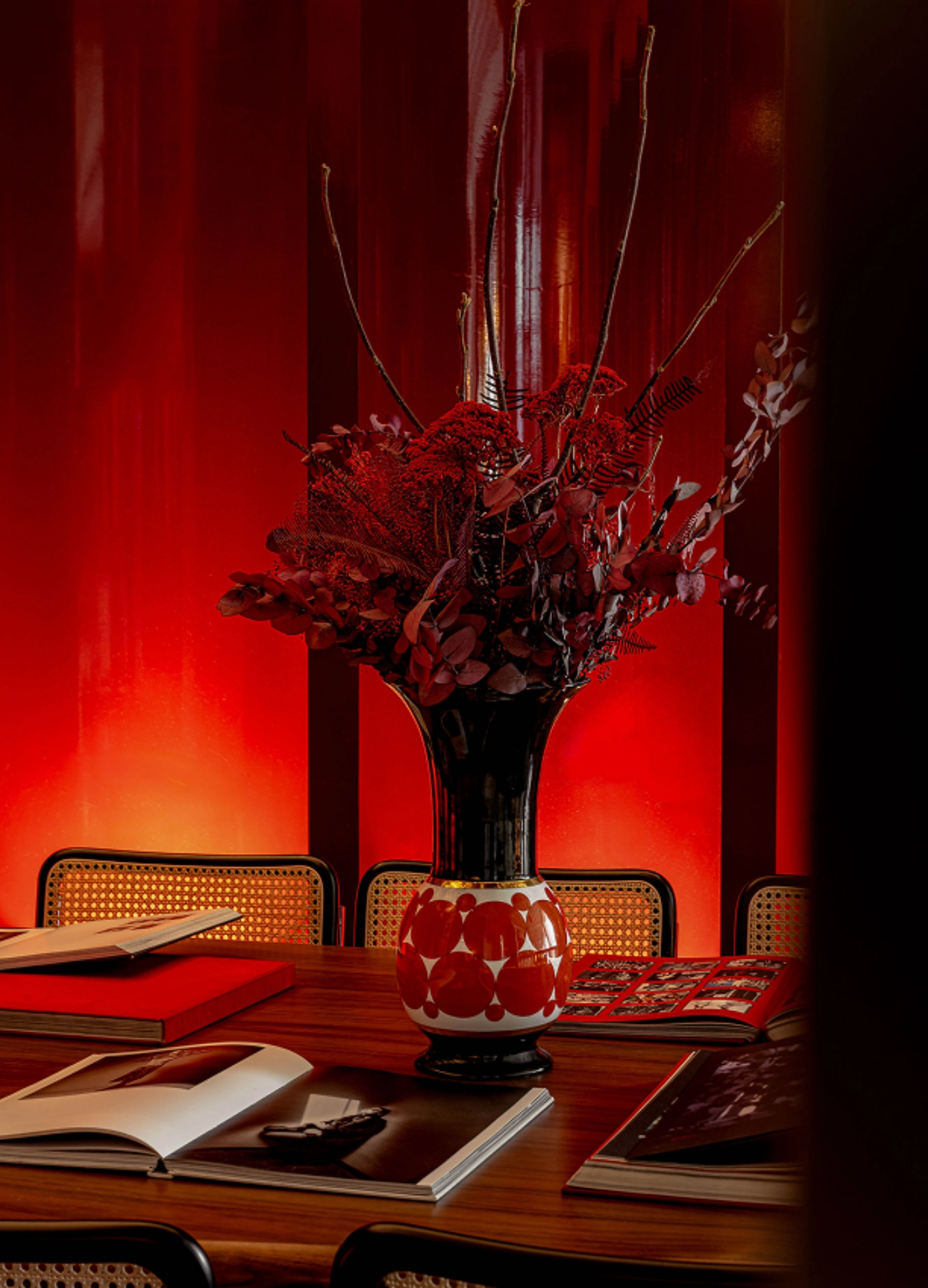 Jarrón decorativo alto con flores y ramas rojas sobre una mesa de madera rodeada de sillas, con álbumes de fotos abiertos bajo una cálida iluminación roja.