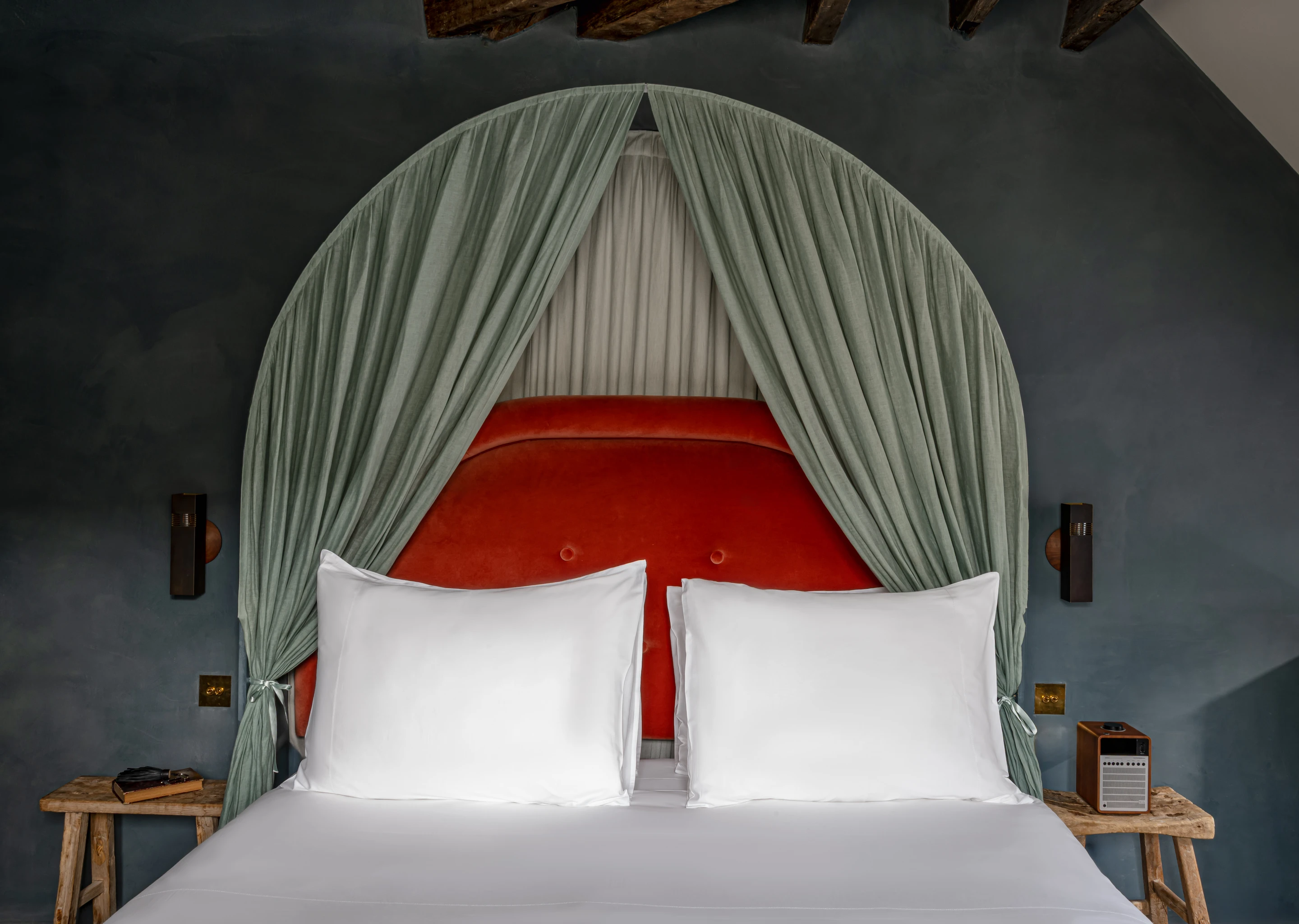 Bed with a red upholstered headboard framed by green draped curtains and two white pillows, flanked by wooden bedside tables with a vintage radio and a book.