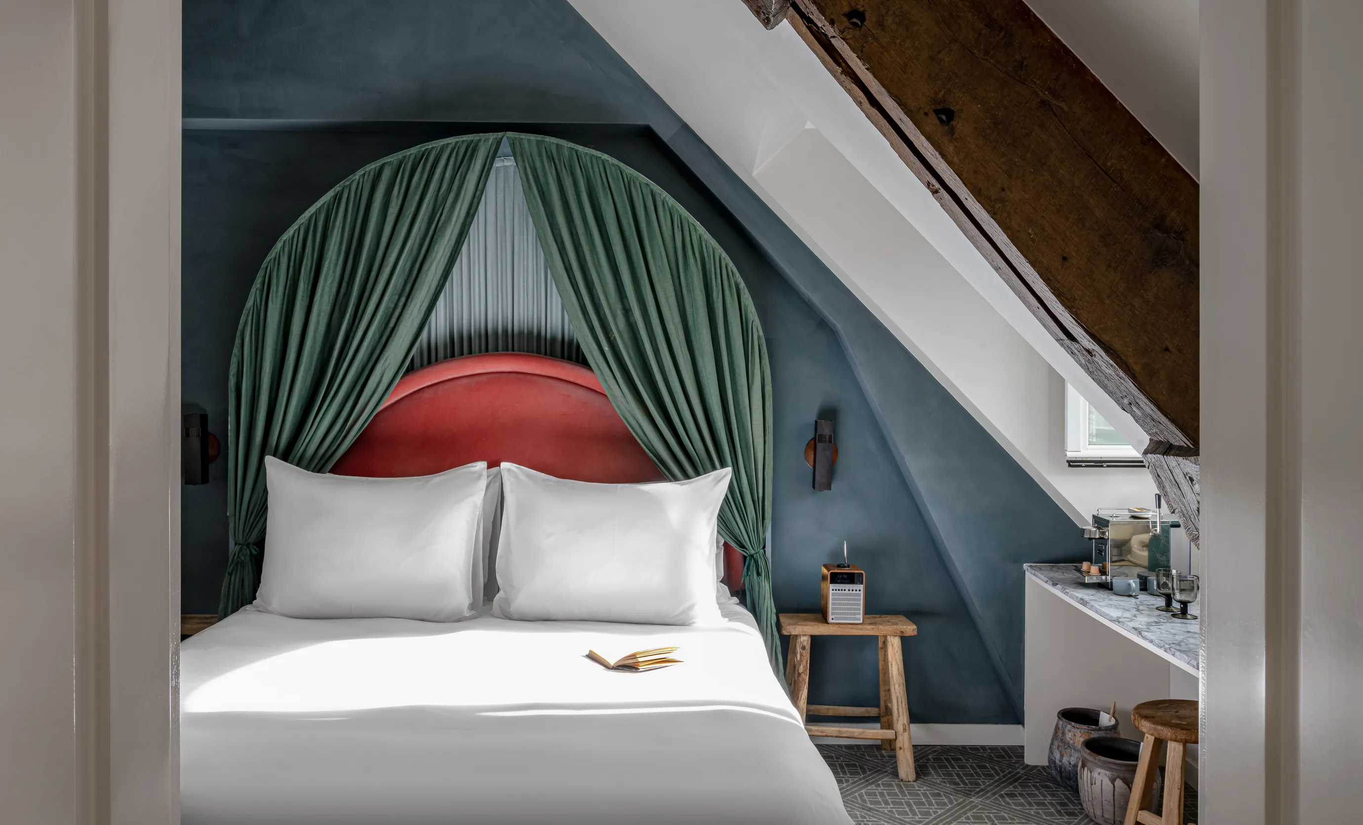 Cozy bedroom with white bed linens, red headboard framed by green curtains, rustic wooden stool, radio, and marble countertop under slanted ceiling.