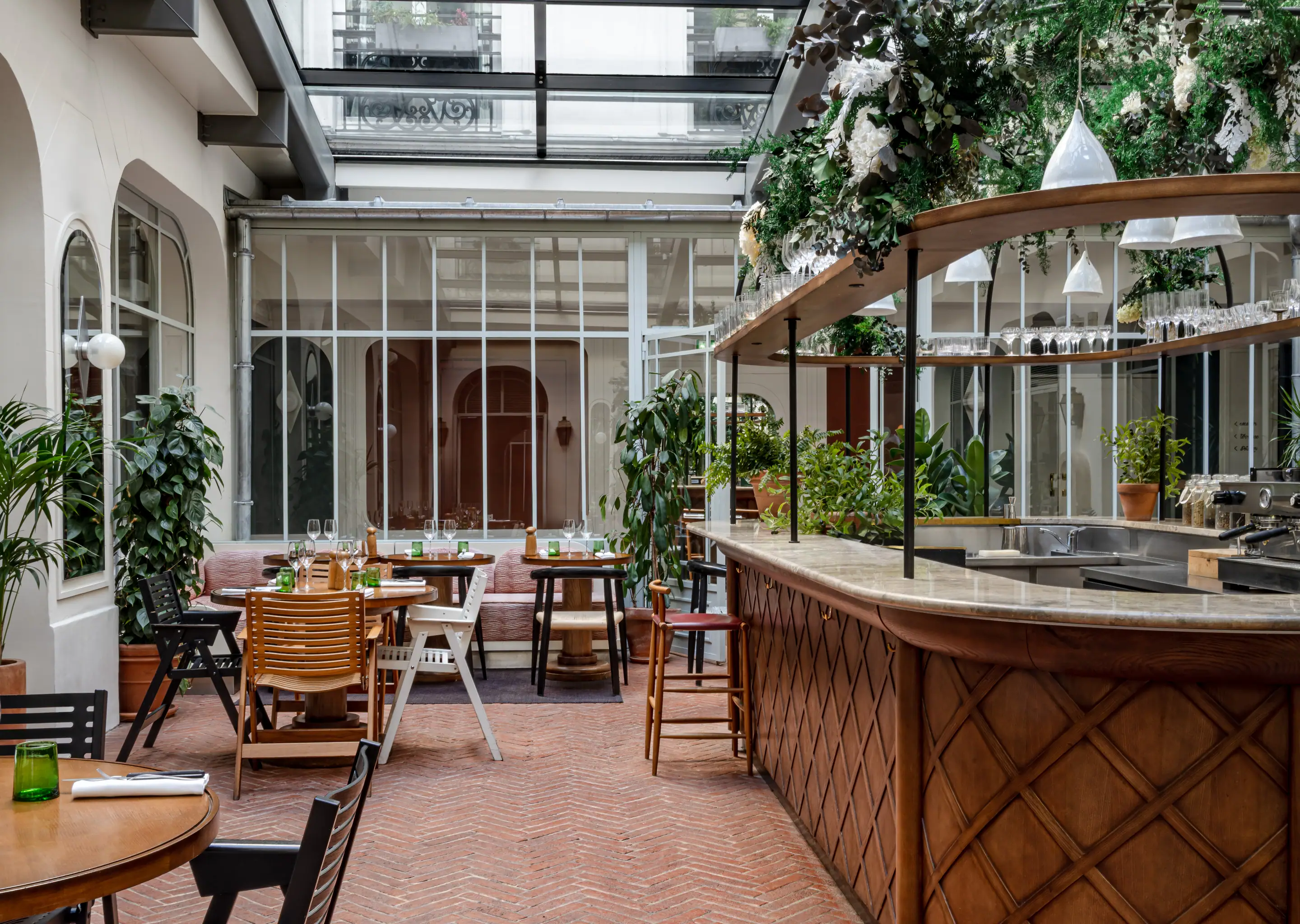 Indoor courtyard restaurant with wooden tables and chairs, a marble-topped bar adorned with greenery and hanging glass lights, under a glass ceiling.