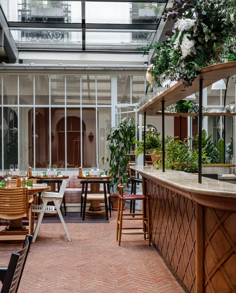 Indoor restaurant seating area with wooden tables, various chairs, a bar with floral decorations, and large windows under a glass ceiling.