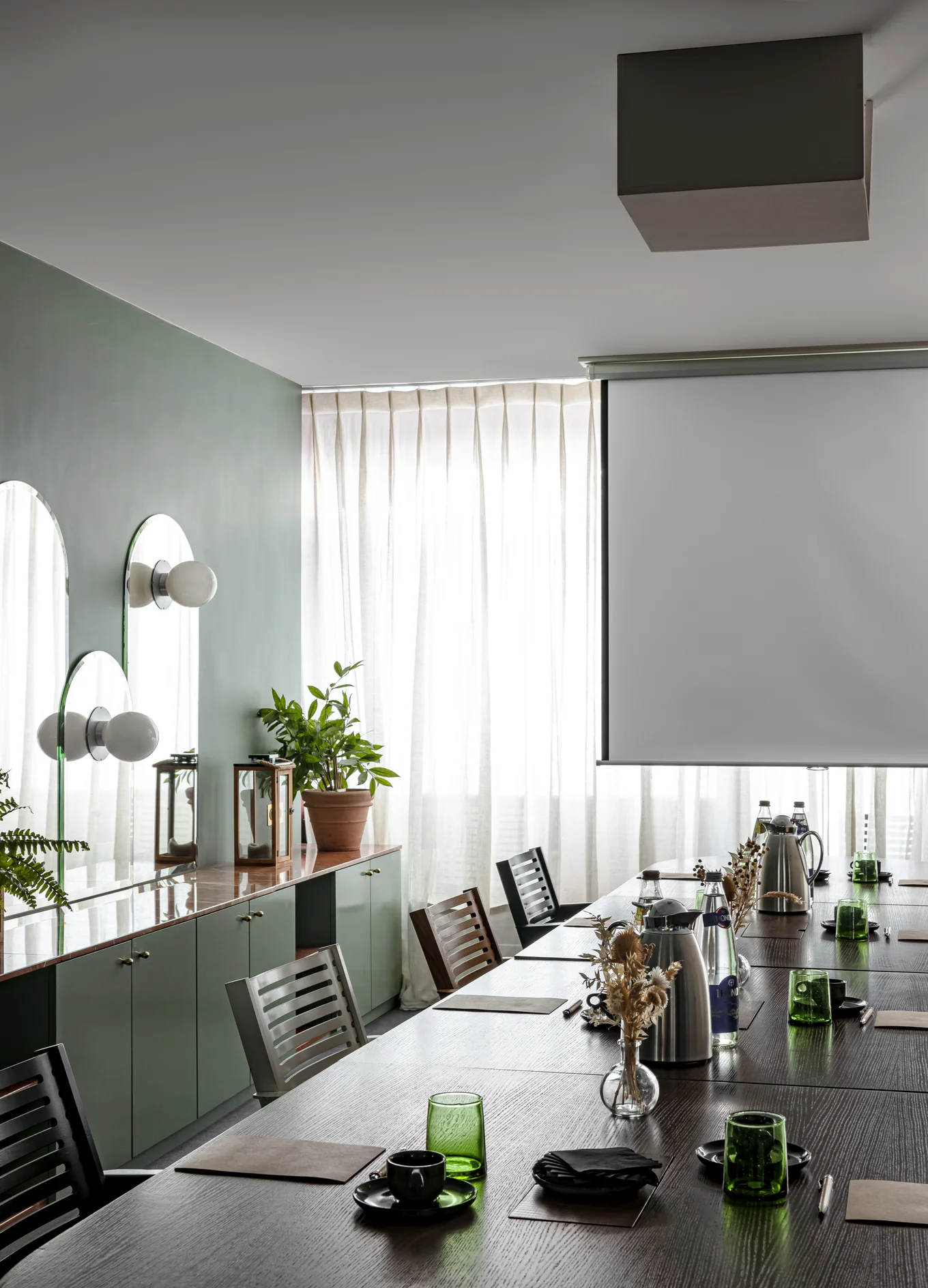 Conference room with a long wooden table set with green glasses, coffee cups, notebooks, and water bottles, featuring a large projection screen and sideboard with plants against a window with sheer white curtains.