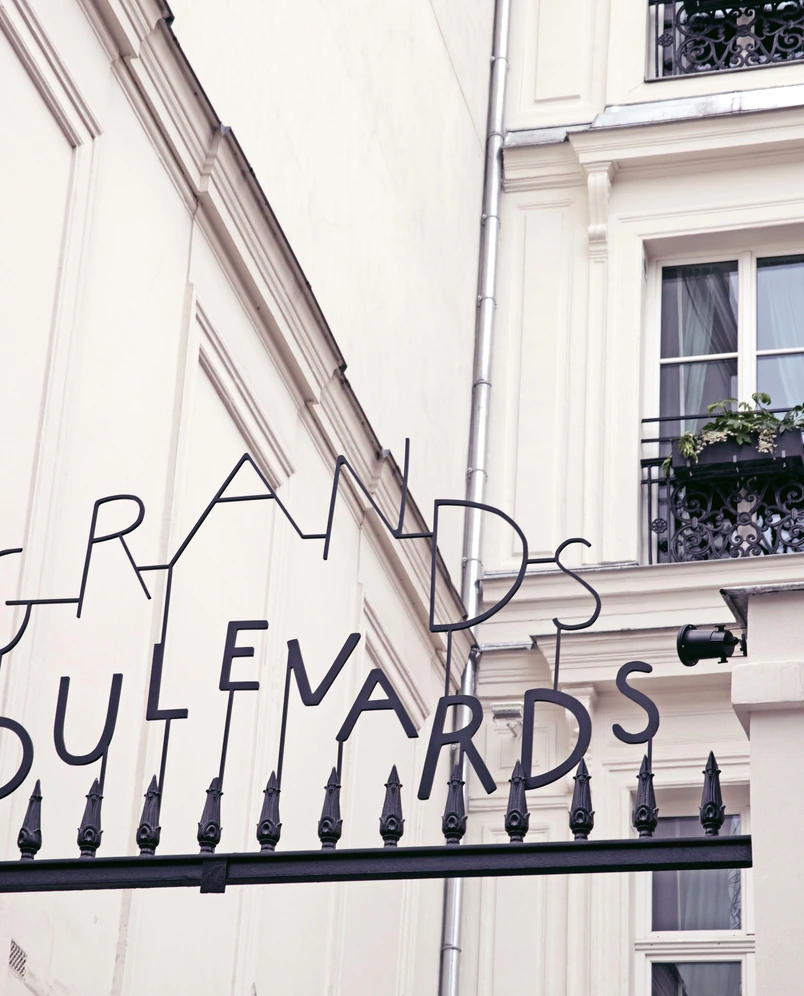 Black metal sign reading 'Grands Boulevards' mounted on black iron fence in front of a white classical building with windows and a flowered balcony.