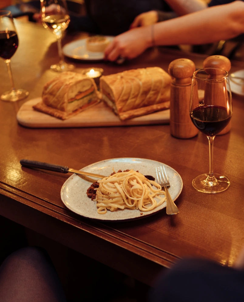 Plate of pasta with fork and knife, glass of red wine, and sliced pastry on a wooden table in a restaurant setting.