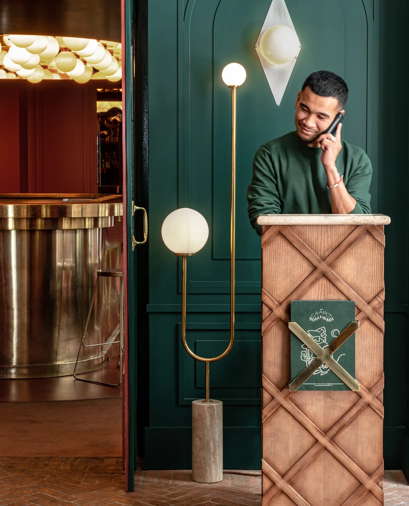 Hotel receptionist standing behind wooden podium with green menu, talking on a phone in a stylish lobby with green walls and modern lamps.