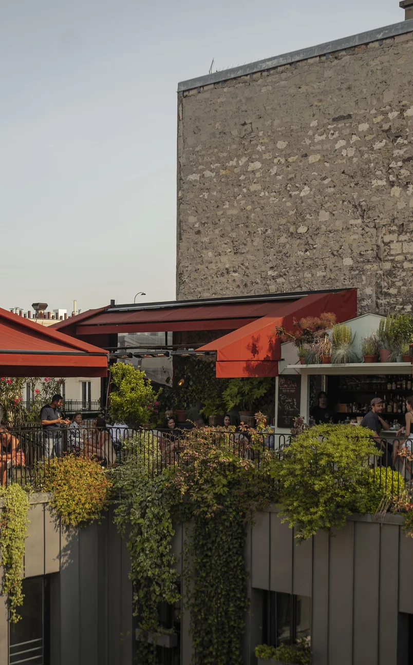 Terrace bar with red awnings, green plants, and people socializing on a rooftop with a stone wall background.
