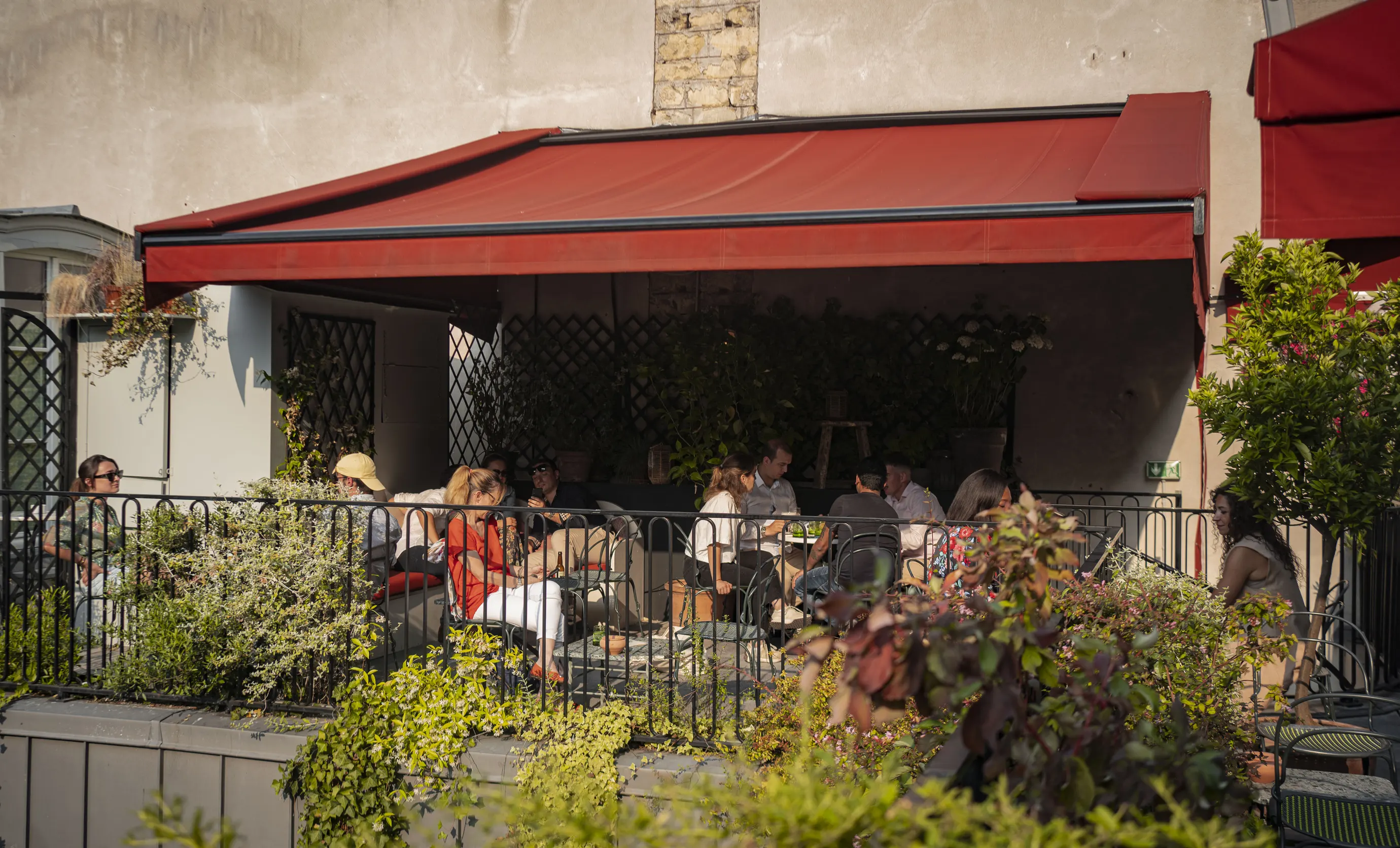Group of people sitting and talking under a red awning at an outdoor café with plants around.