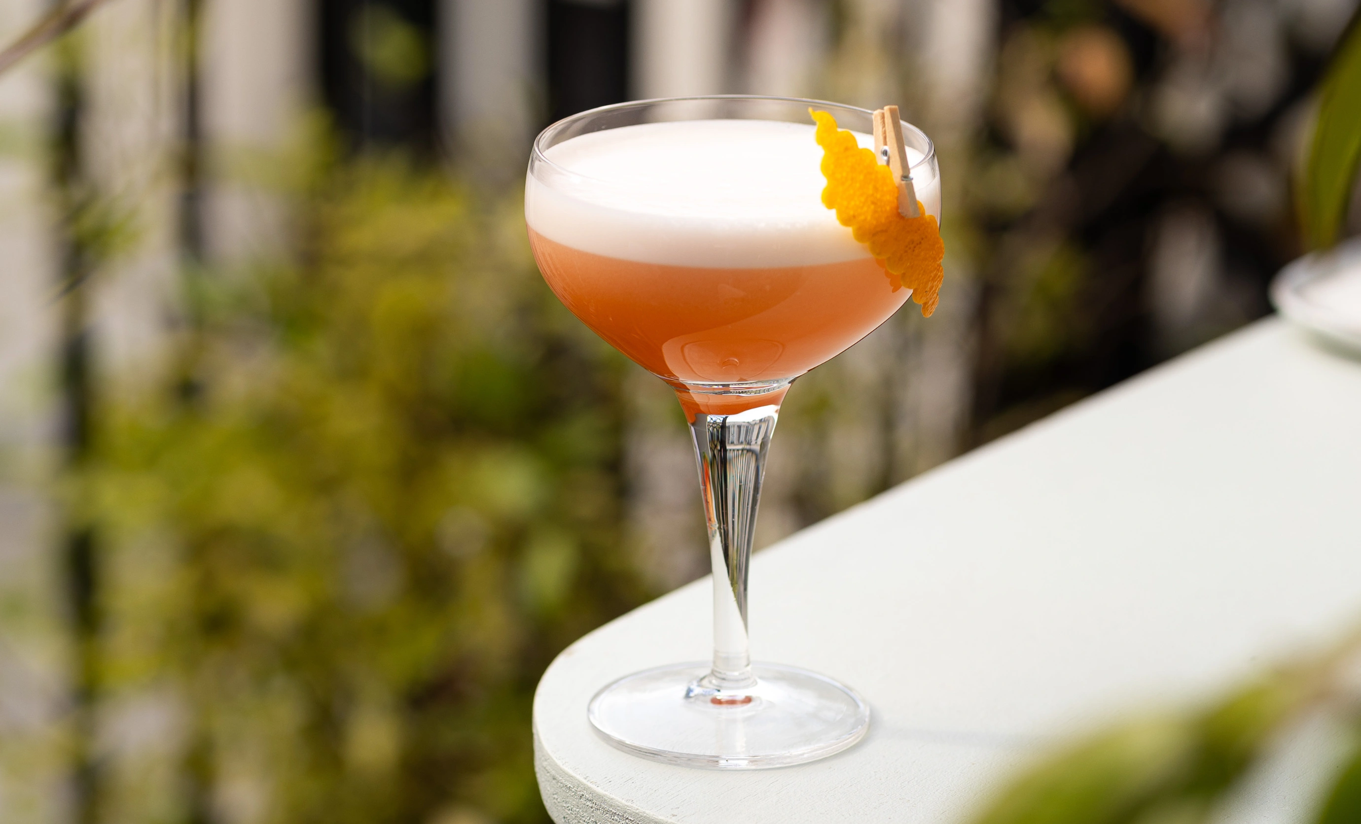 Cocktail glass with a frothy peach-colored drink garnished with a twisted orange peel clipped to the rim.