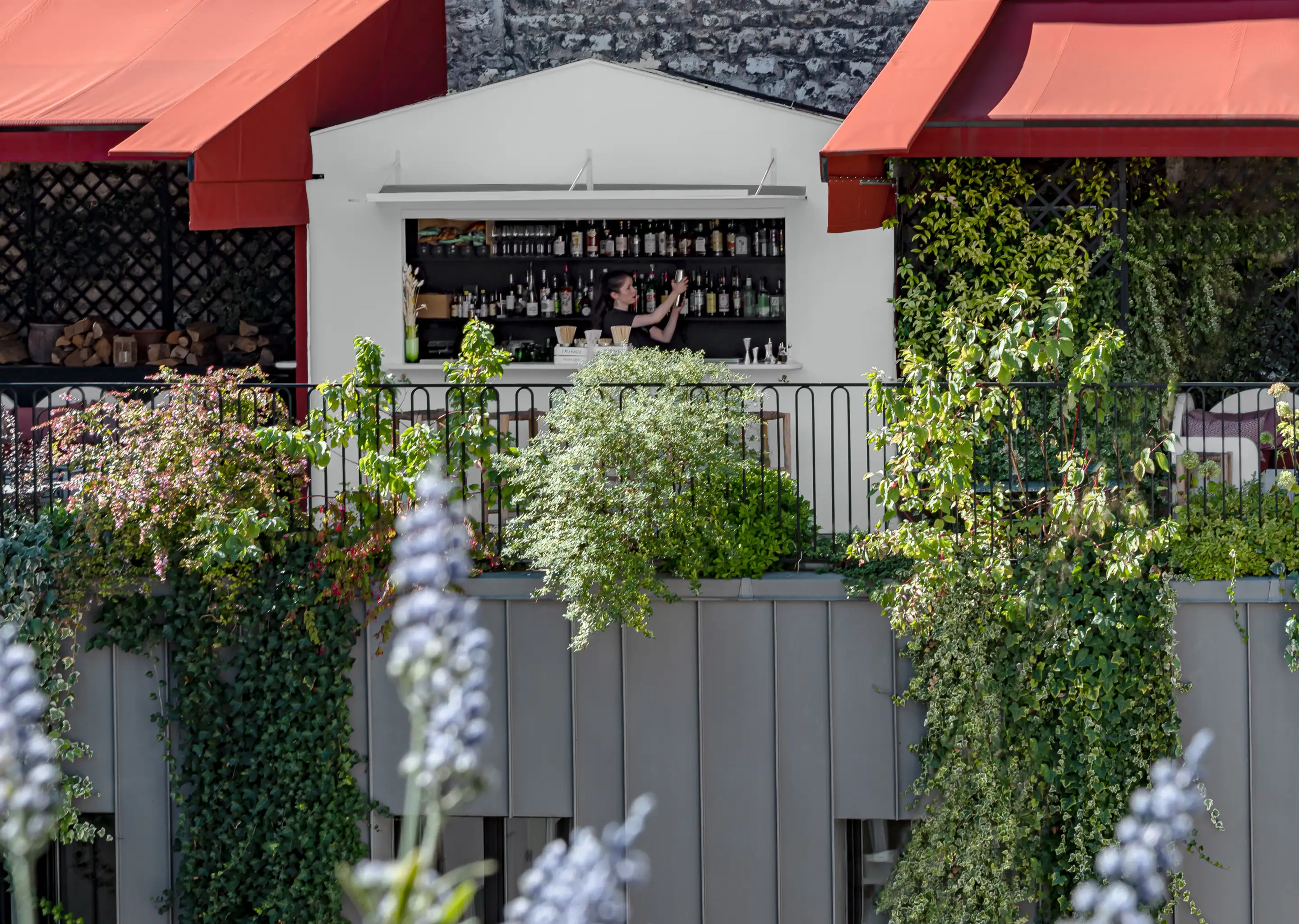 Outdoor bar with a woman arranging bottles behind the counter, surrounded by greenery and red awnings.