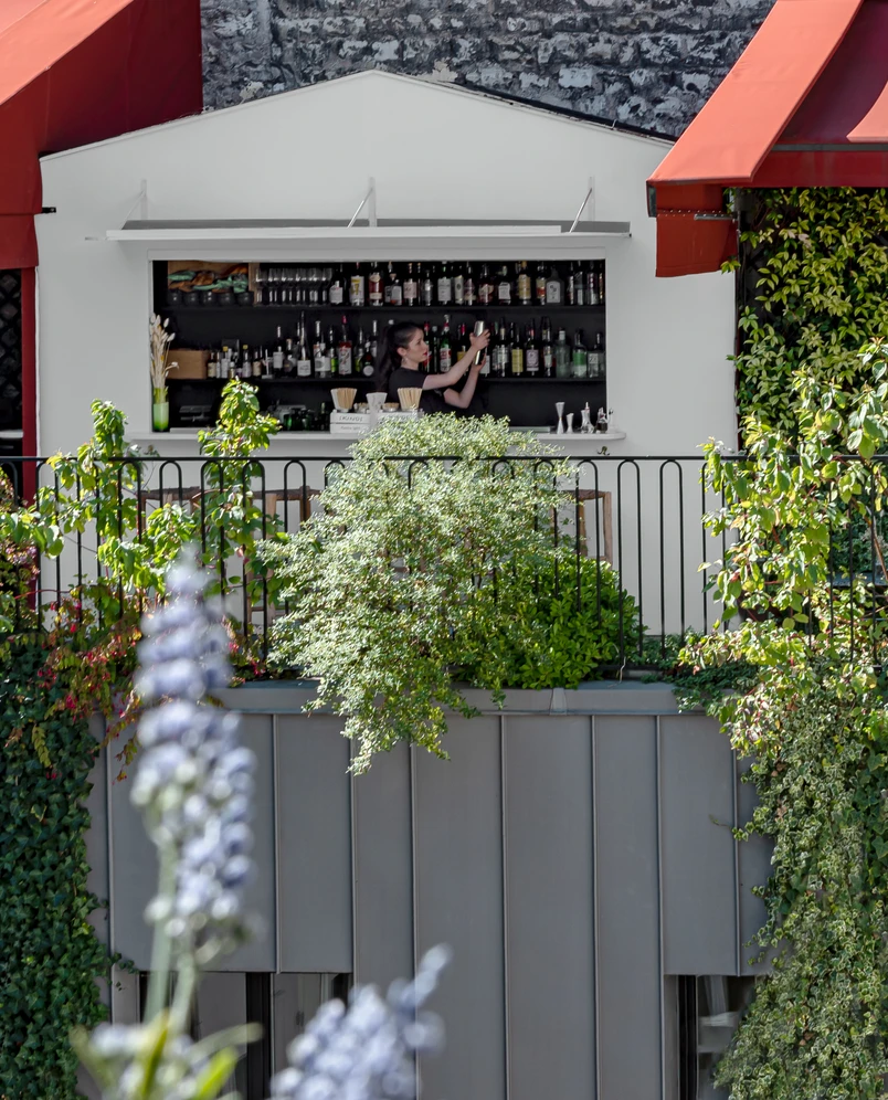Bartender arranging bottles in an outdoor bar window surrounded by greenery and red awnings.
