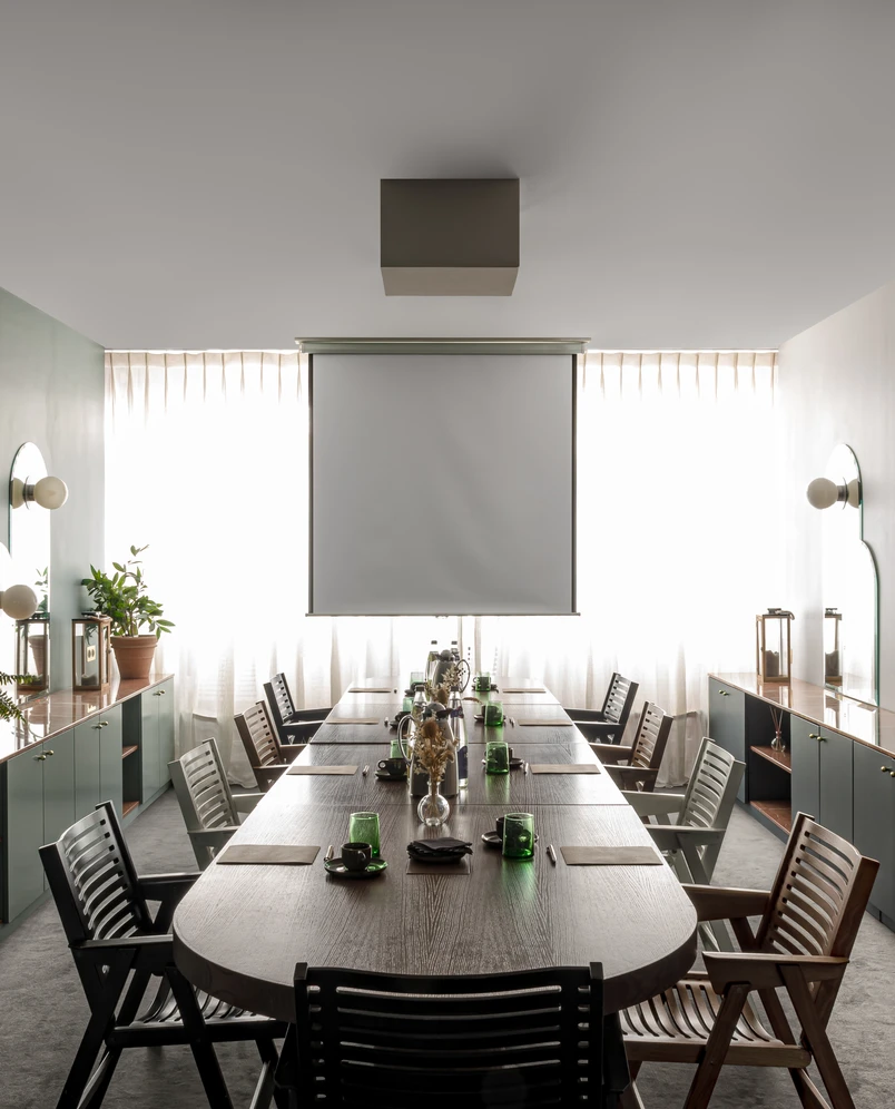Sala de conferências moderna com uma longa mesa de madeira, cadeiras mistas, copos verdes e uma tela de projetor em frente a cortinas brilhantes.
