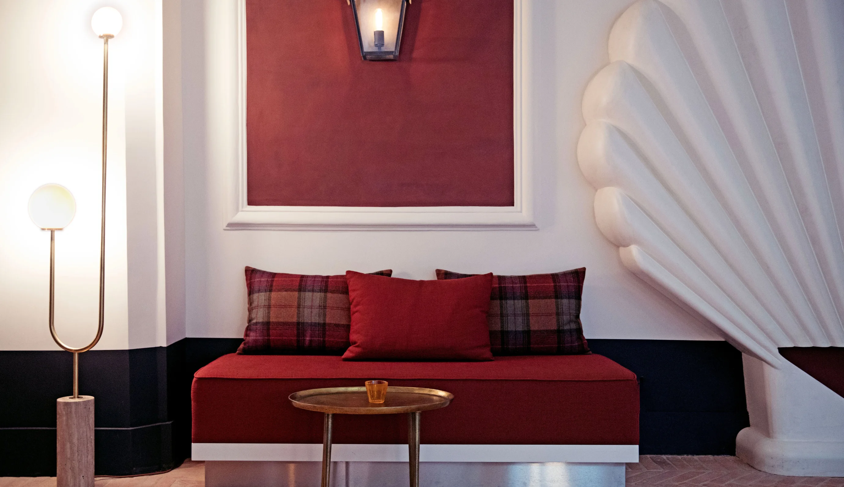 Modern seating area with a red cushioned bench, plaid and solid pillows, brass round table, and decorative white wall panel.