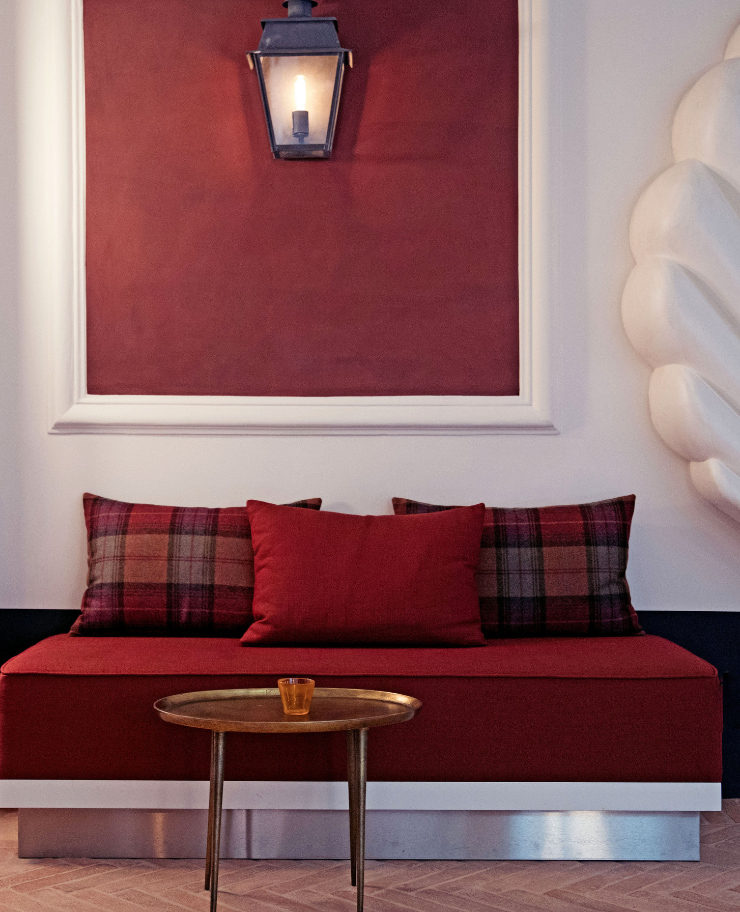 Cozy seating area with a red cushioned bench, plaid and solid red pillows, a small round brass table with an amber glass, and a lit lantern on a red framed wall.