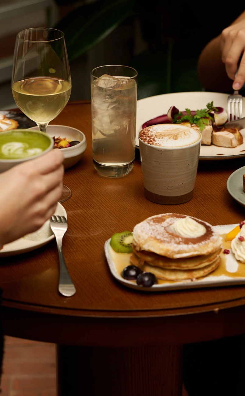 Table with pancakes topped with cream and fruit, latte in a ceramic cup, glass of iced water, white wine, and a hand holding a matcha latte.