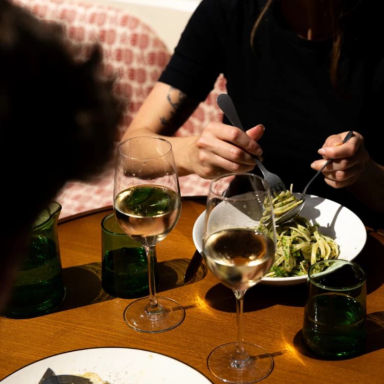 Person twirling green pasta with utensils at a wooden table set with two wine glasses filled with rosé wine and green glassware.
