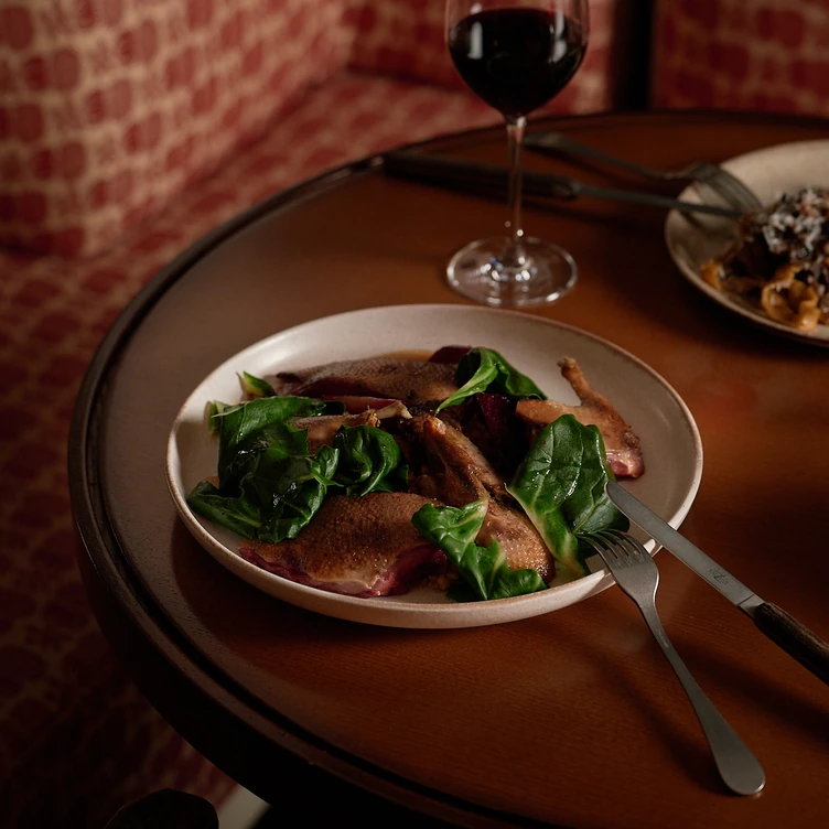 Plate with cooked duck breast pieces and leafy greens on a wooden table with a fork, knife, and a glass of red wine.