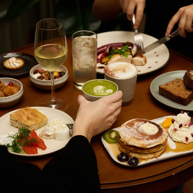 Table with a variety of brunch dishes including pancakes with fruit and cream, bread with cheese and smoked salmon, pastries, a glass of white wine, iced water, a matcha latte, and a cappuccino, with two people dining.