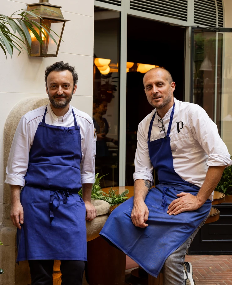 Two male chefs wearing white shirts and blue aprons sitting and standing outside a restaurant near a table with plants.