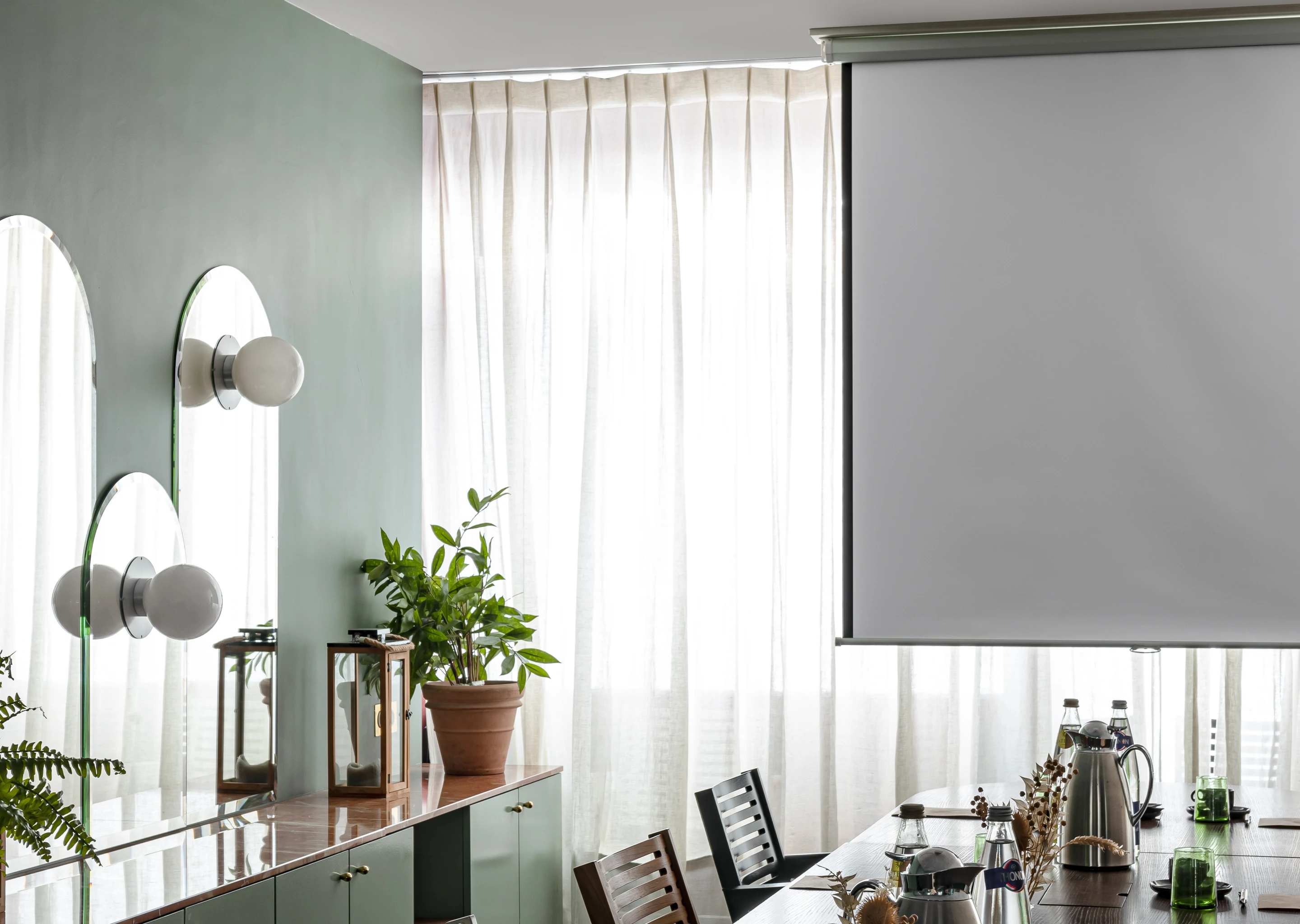 Sala de reuniones luminosa con una mesa de conferencias, sillas, plantas y una pantalla de proyección en blanco frente a cortinas transparentes.