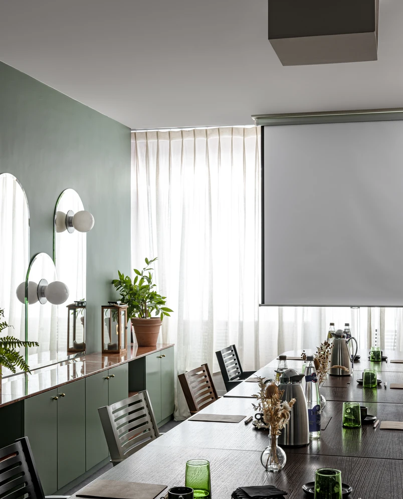 Moderna sala de conferencias con una mesa larga de madera oscura con vasos verdes, jarras de agua de metal y arreglos florales secos cerca de una ventana grande con cortinas transparentes y una pantalla de proyección.