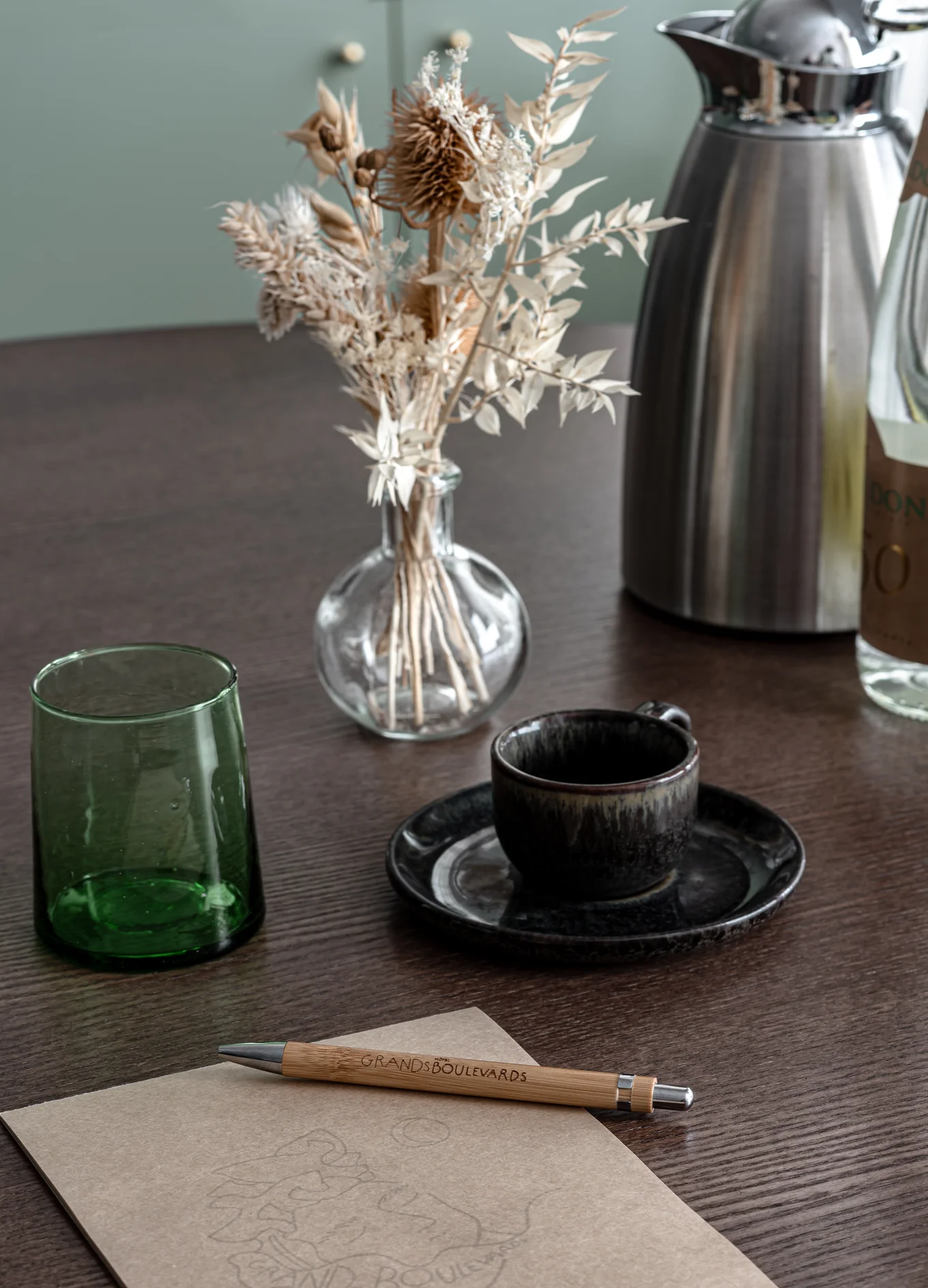Mesa de madera oscura con un jarrón de vidrio con flores secas, un vaso verde, una taza de cerámica negra con platillo, una cafetera de metal, una botella y un cuaderno con un bolígrafo de madera con la etiqueta «GRANDS BOULEVARDS».