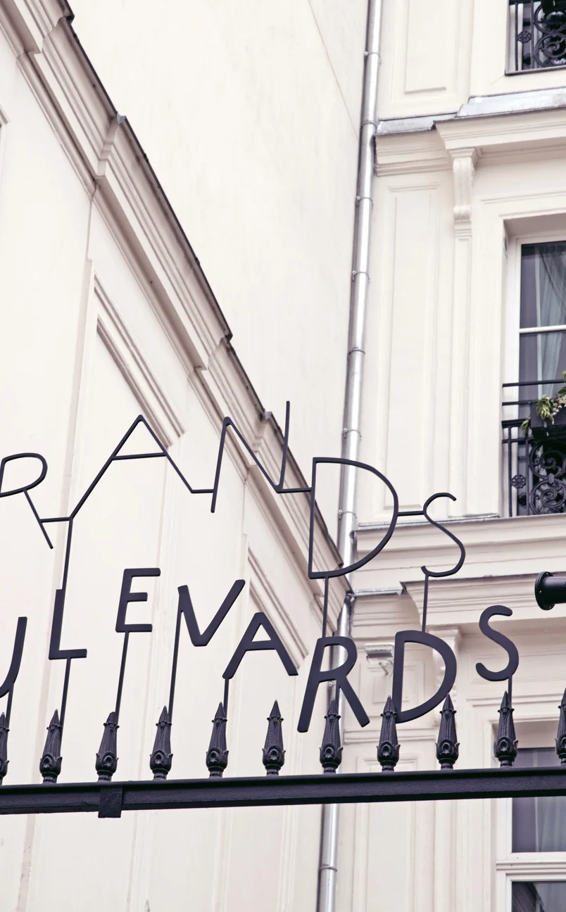 Letras metálicas negras que deletrean «GRANDS BOULEVARDS» en una valla decorativa frente a la fachada de un edificio de color beige.
