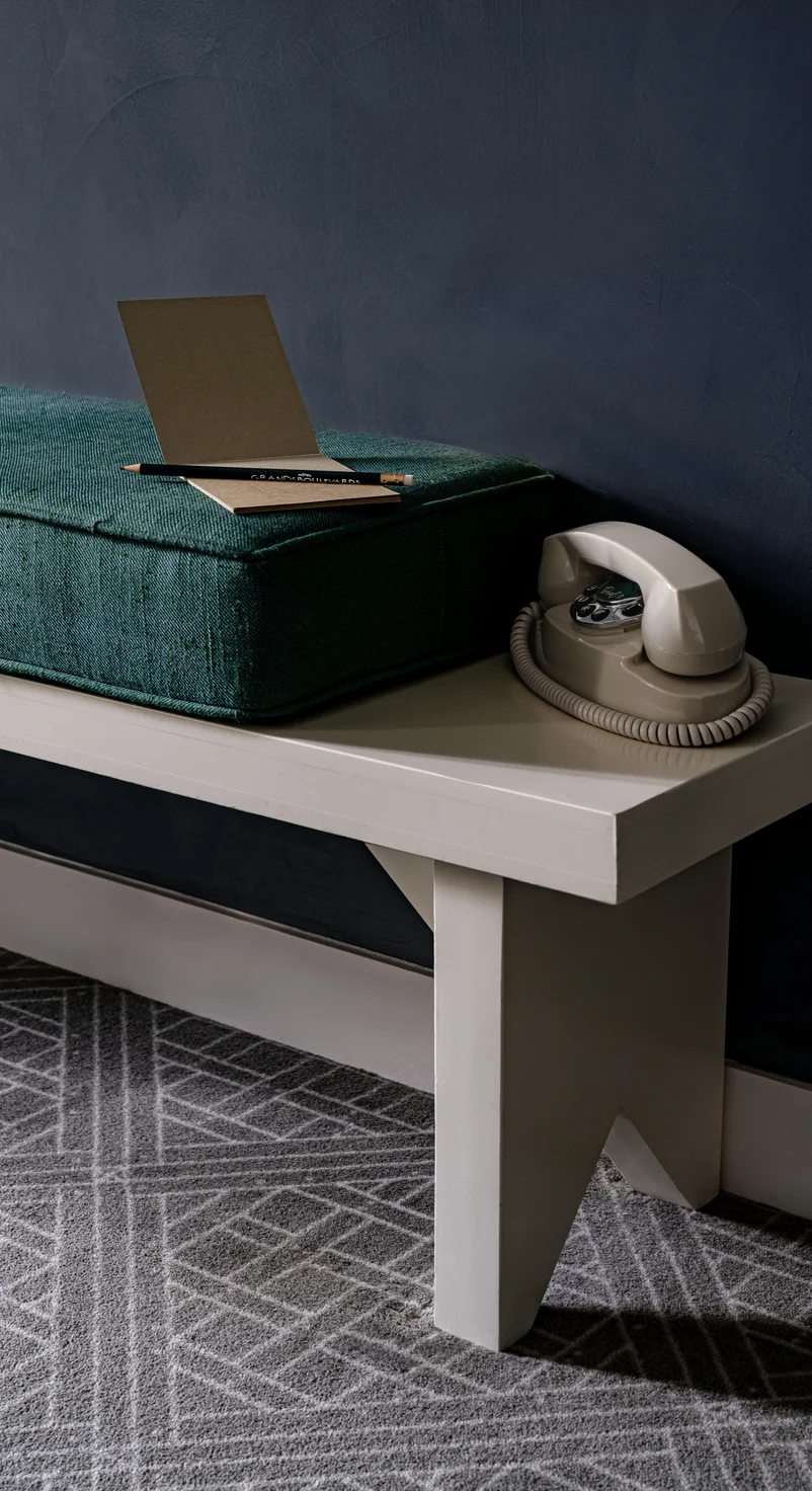 Teléfono con esfera giratoria de color beige y cojín verde con lápiz y bloc de notas sobre un banco blanco contra una pared azul oscuro.
