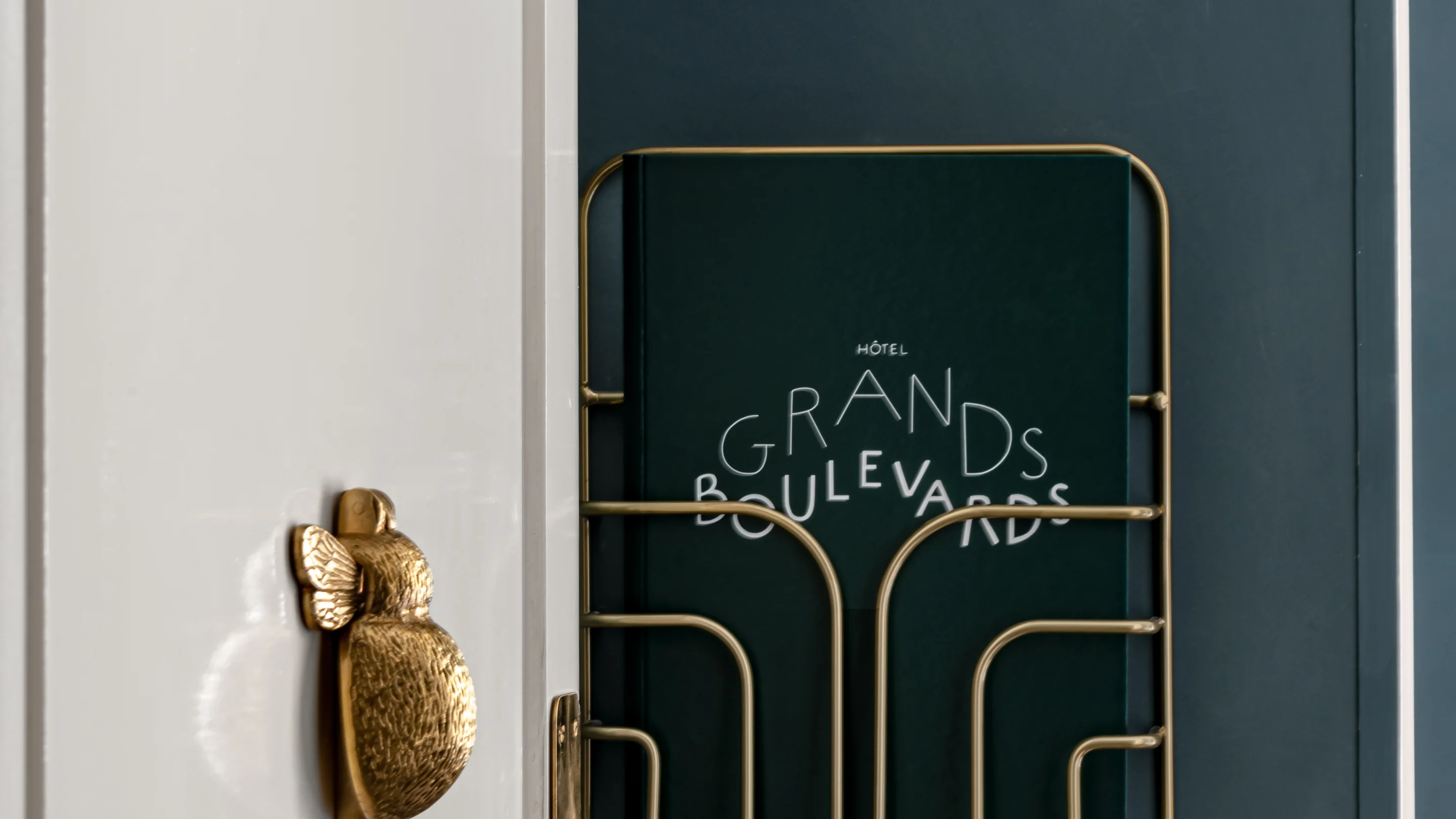 Green book titled 'Hôtel Grands Boulevards' held in a golden wire rack mounted on a dark wall with a golden bird-shaped door handle nearby.