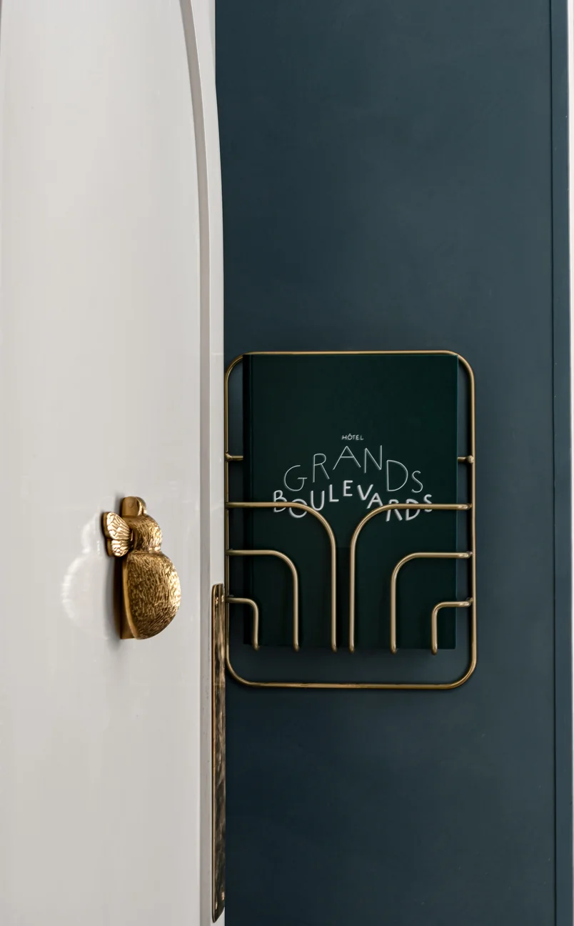 Close-up of a brass bee door handle on a white door next to a dark green sign holder with 'Hôtel Grands Boulevards' text on a dark wall.
