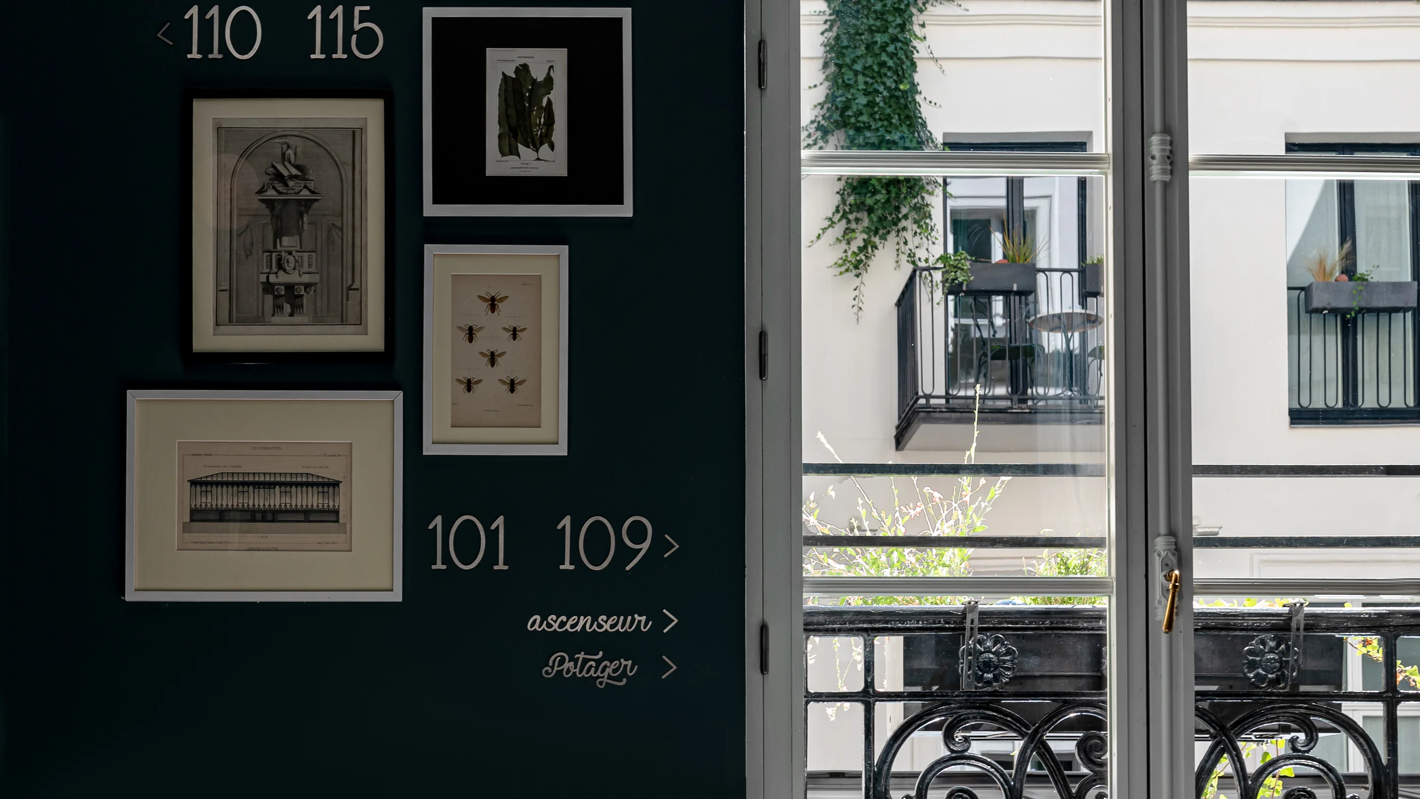 Pared oscura con grabados botánicos y arquitectónicos enmarcados, indicaciones sobre los números de las habitaciones y una ventana que da al balcón y al edificio vecino.
