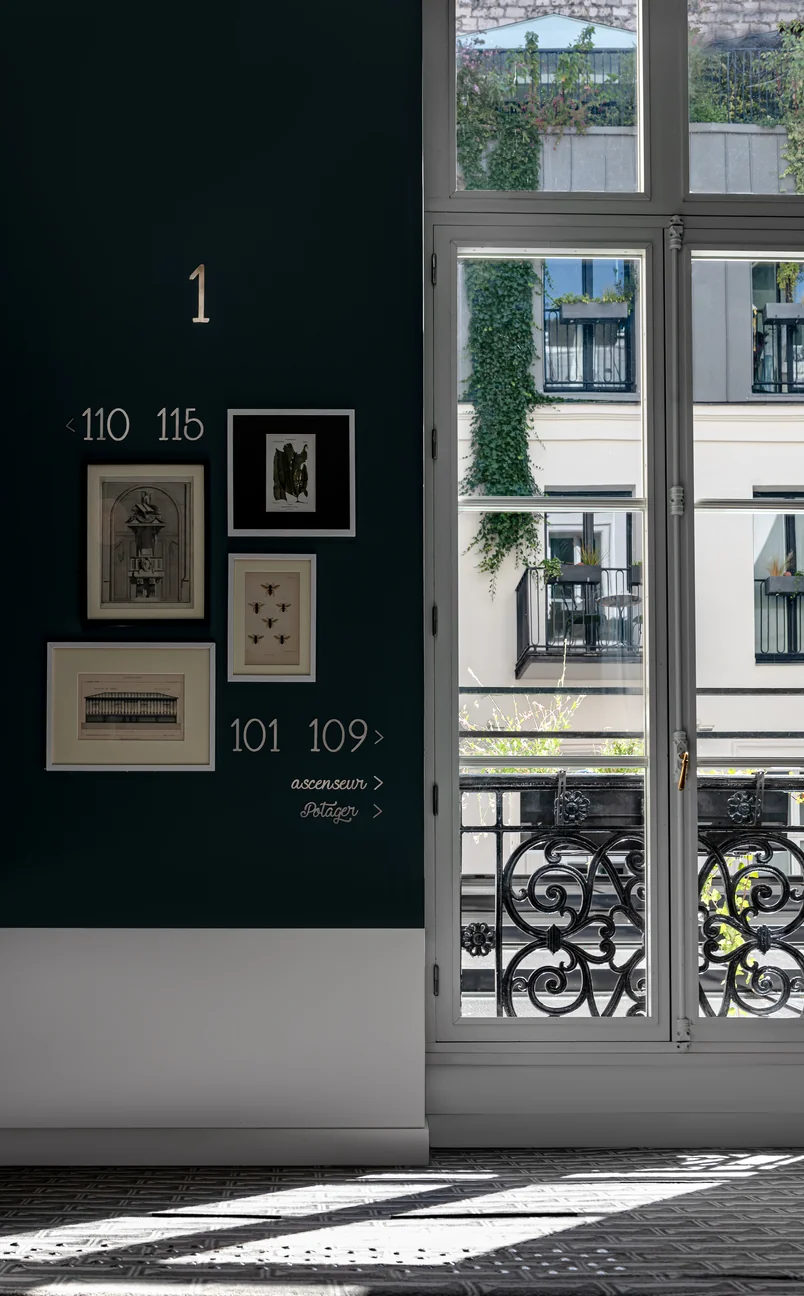 Pasillo interior con pared verde oscuro con obras de arte enmarcadas y direcciones de números de habitación junto a una ventana grande que muestra un balcón y un edificio cubierto de hiedra en el exterior.