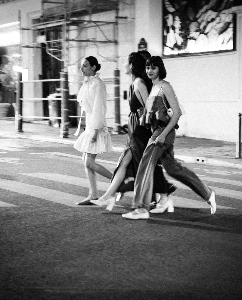 Foto en blanco y negro de cuatro mujeres caminando cogidas del brazo por una calle de la ciudad por la noche, vestidas con elegantes trajes de noche.