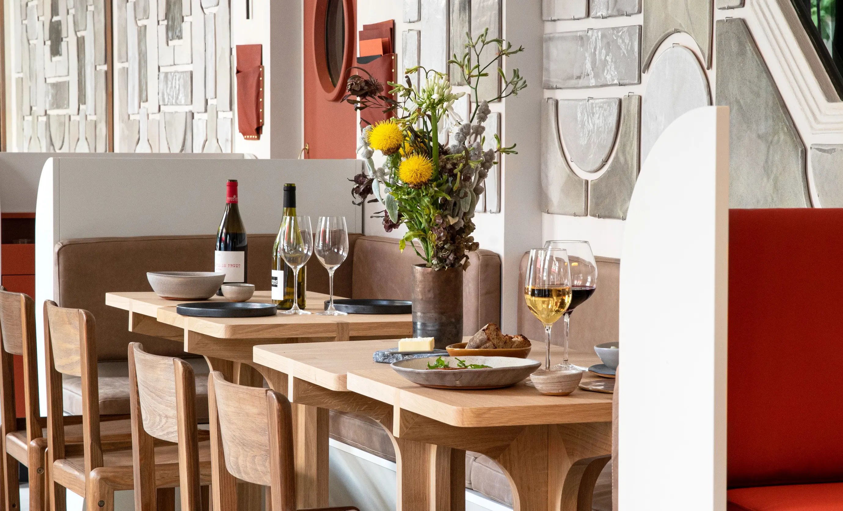 Table set with wine glasses, bottles, plates, bread, and a flower vase in a modern restaurant with wooden chairs and decorative wall panels.