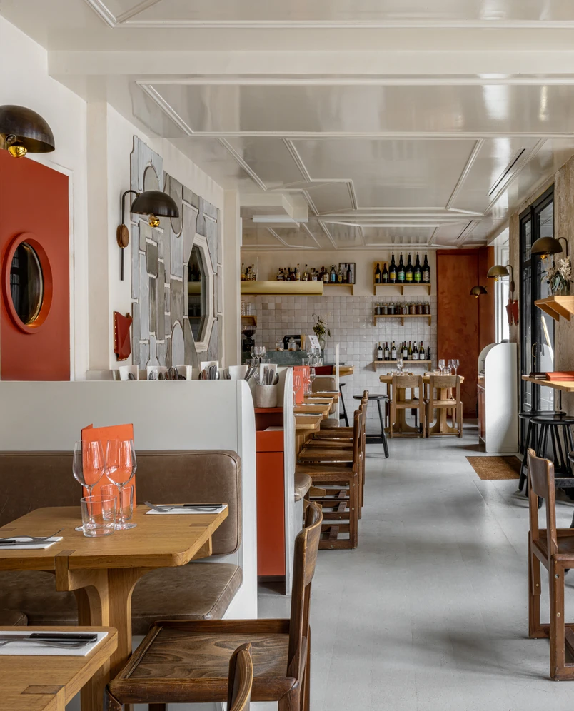 Modern restaurant interior with wooden tables, brown leather chairs and booth seating, and shelves with bottles on the back wall.